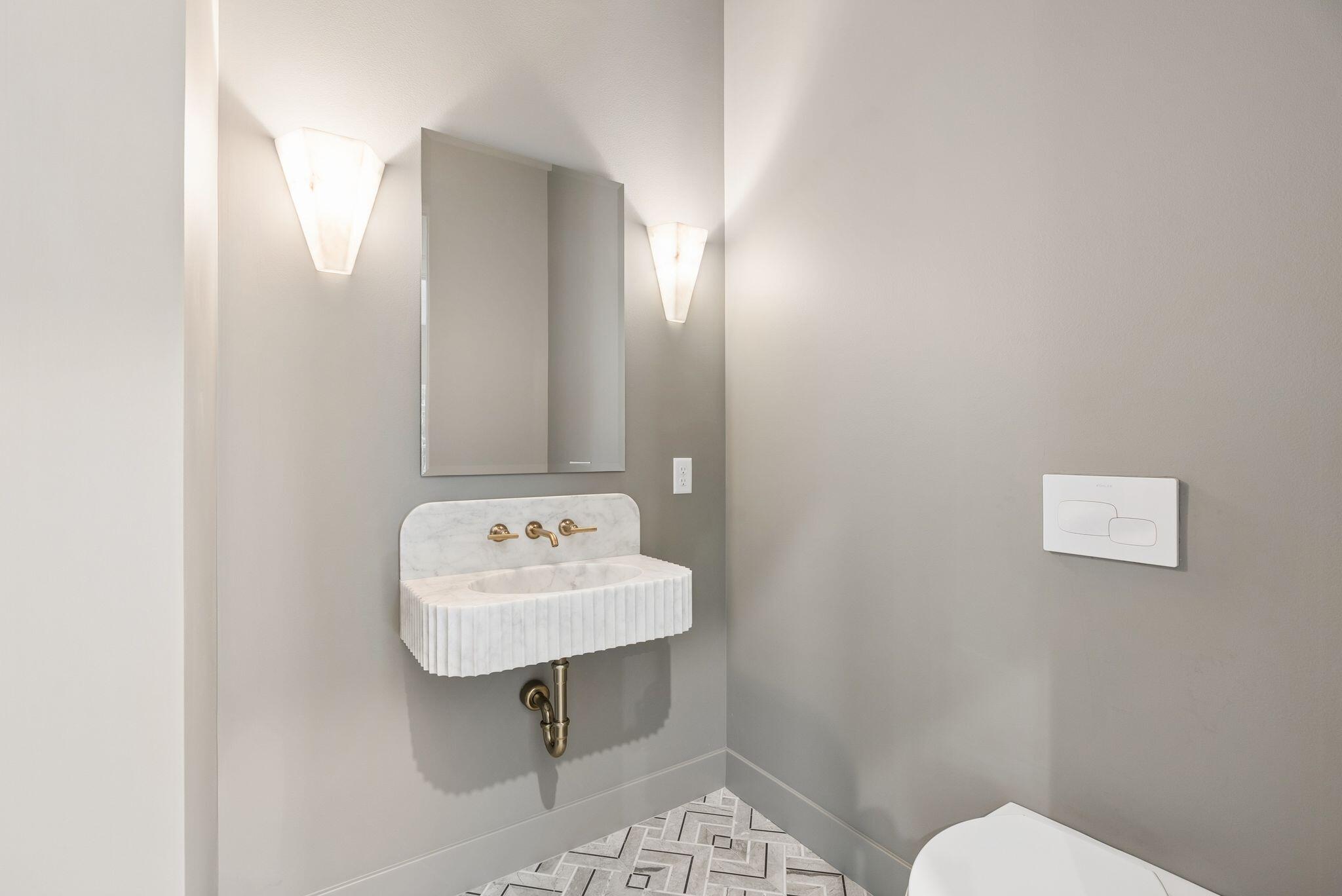 Modern bathroom with a fluted stone sink, brass fixtures, geometric tile floor, and partially visible toilet.