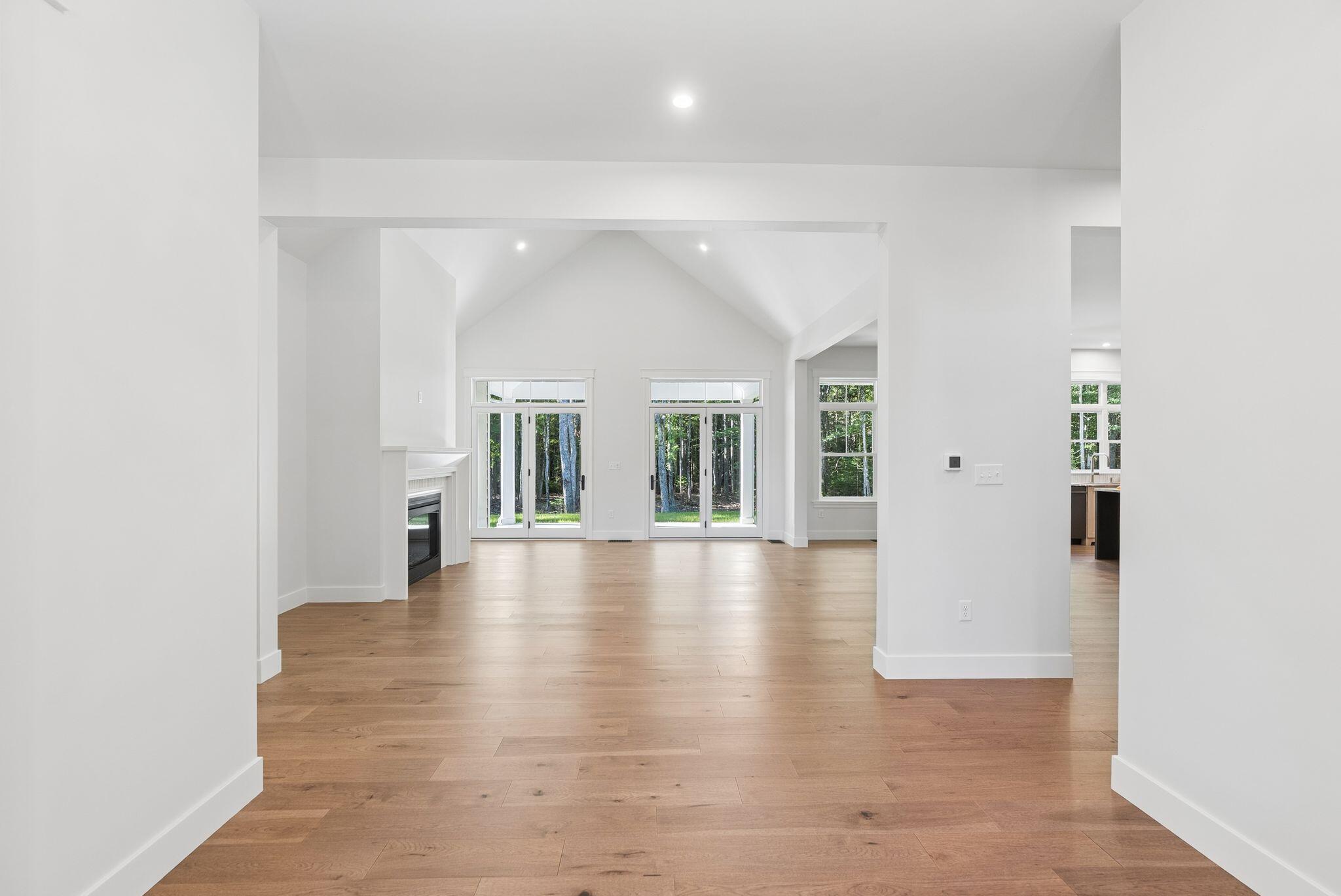 Vacant living area with vaulted ceiling, wood floors, fireplace, and large windows facing trees.