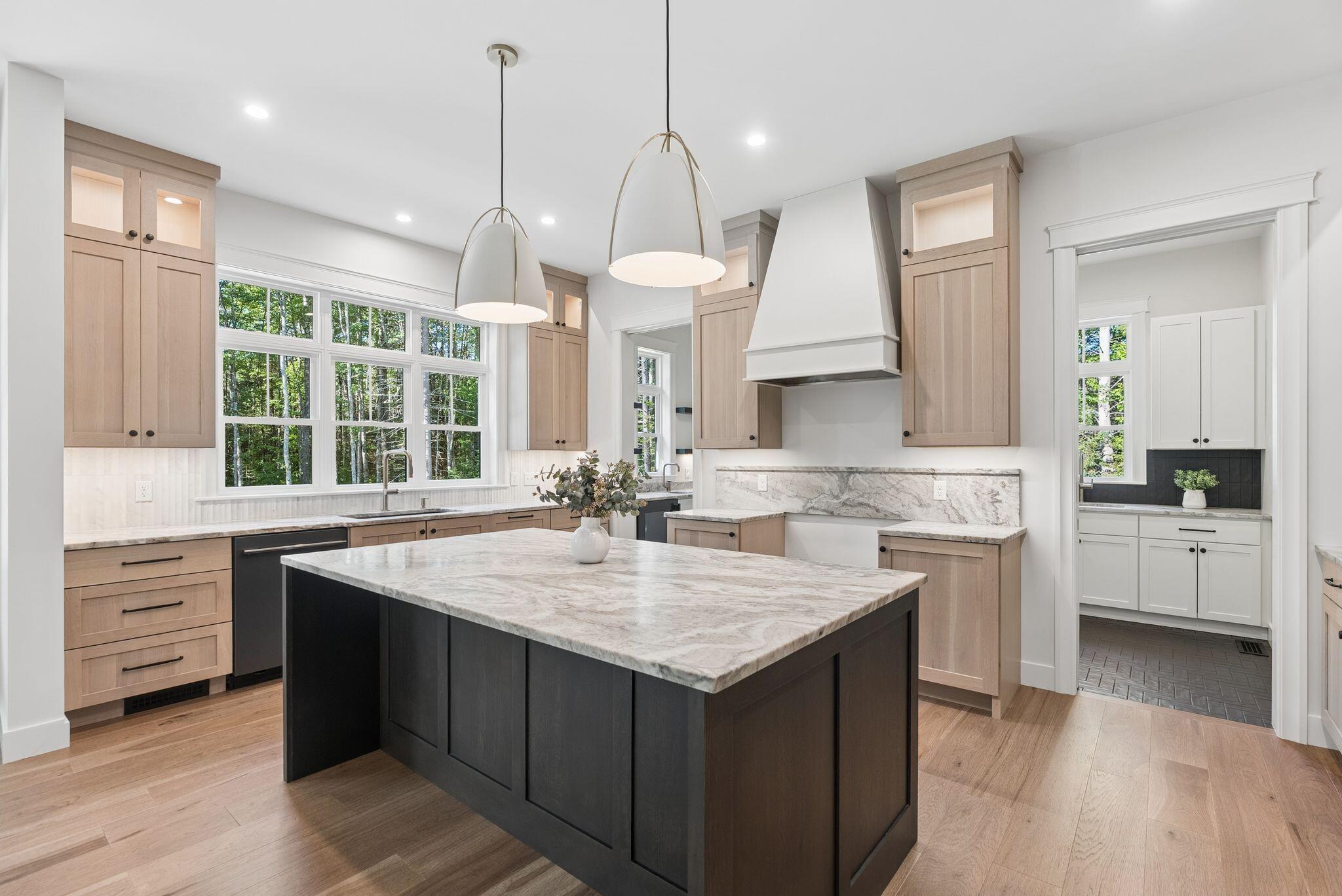 Bright kitchen with a dark stone-topped island, light wood cabinets, and large windows overlooking trees.