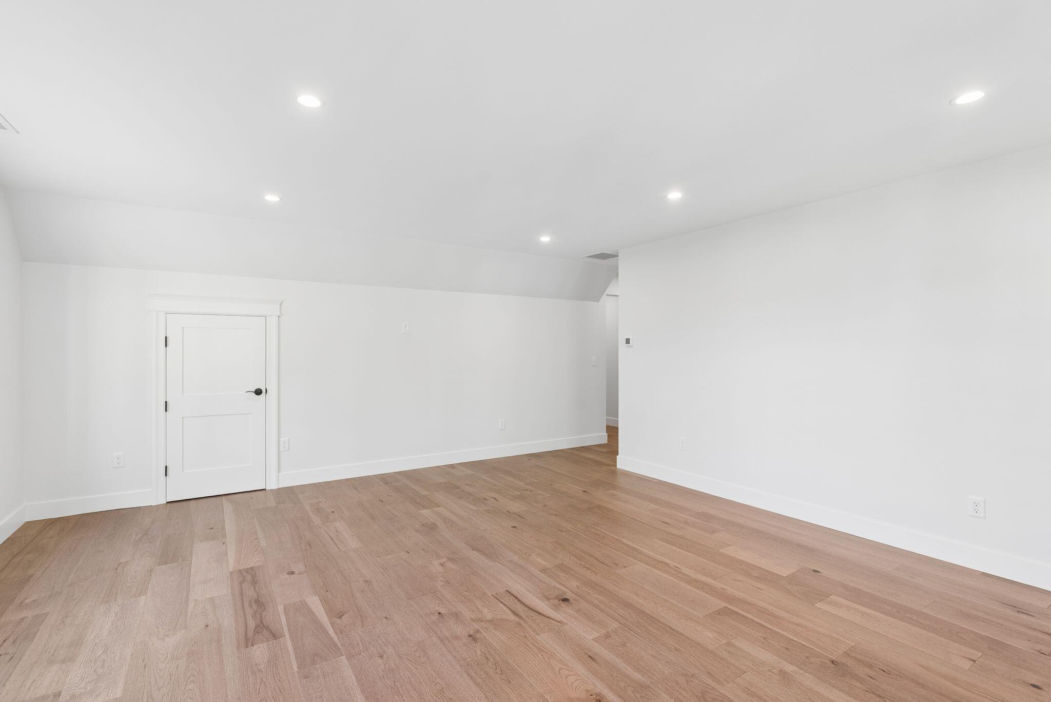 Empty room with light wood floors, white paneled door, and recessed ceiling lights.