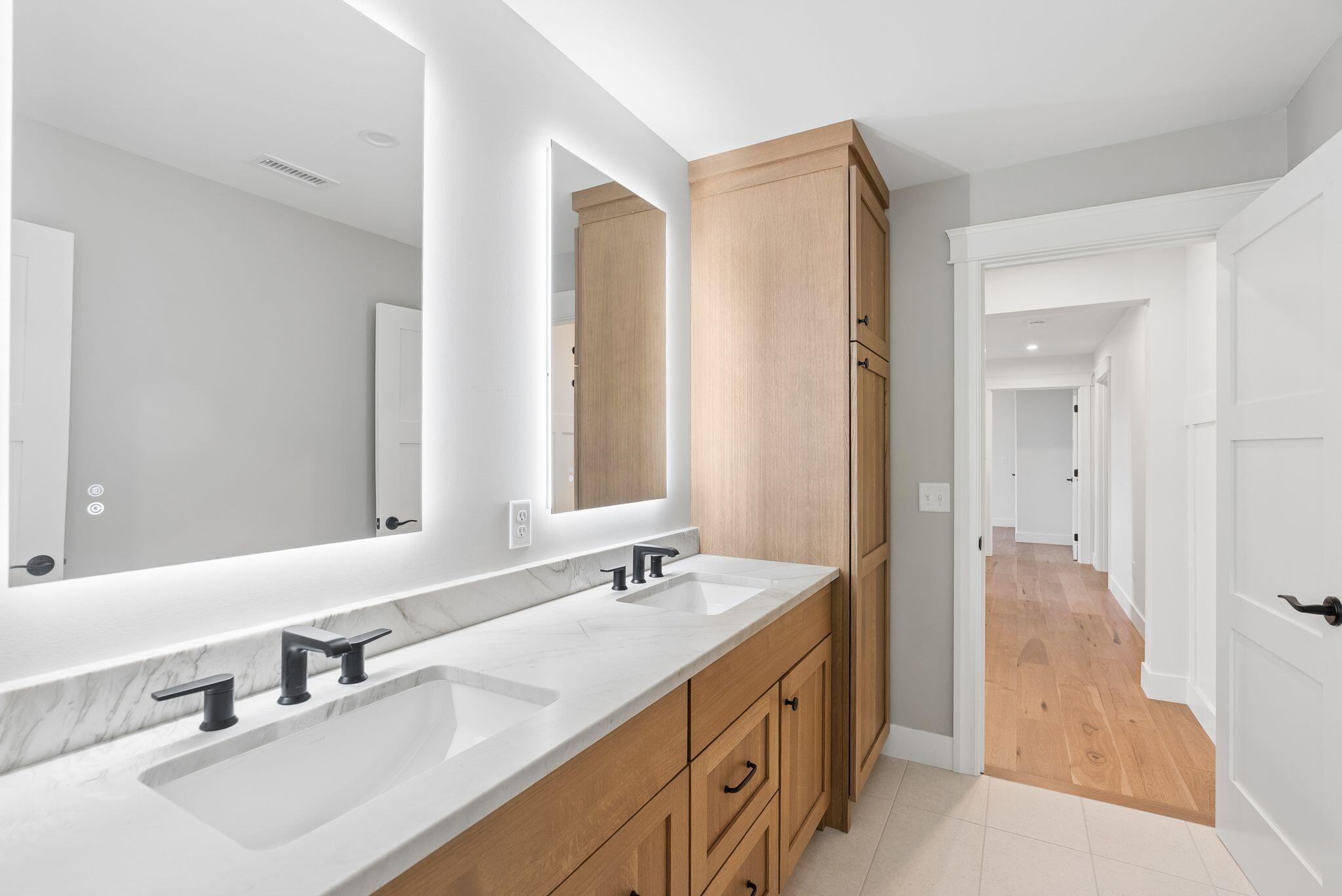 Bathroom with a double-sink vanity, marble-look counters, light wood cabinets, and an illuminated mirror.