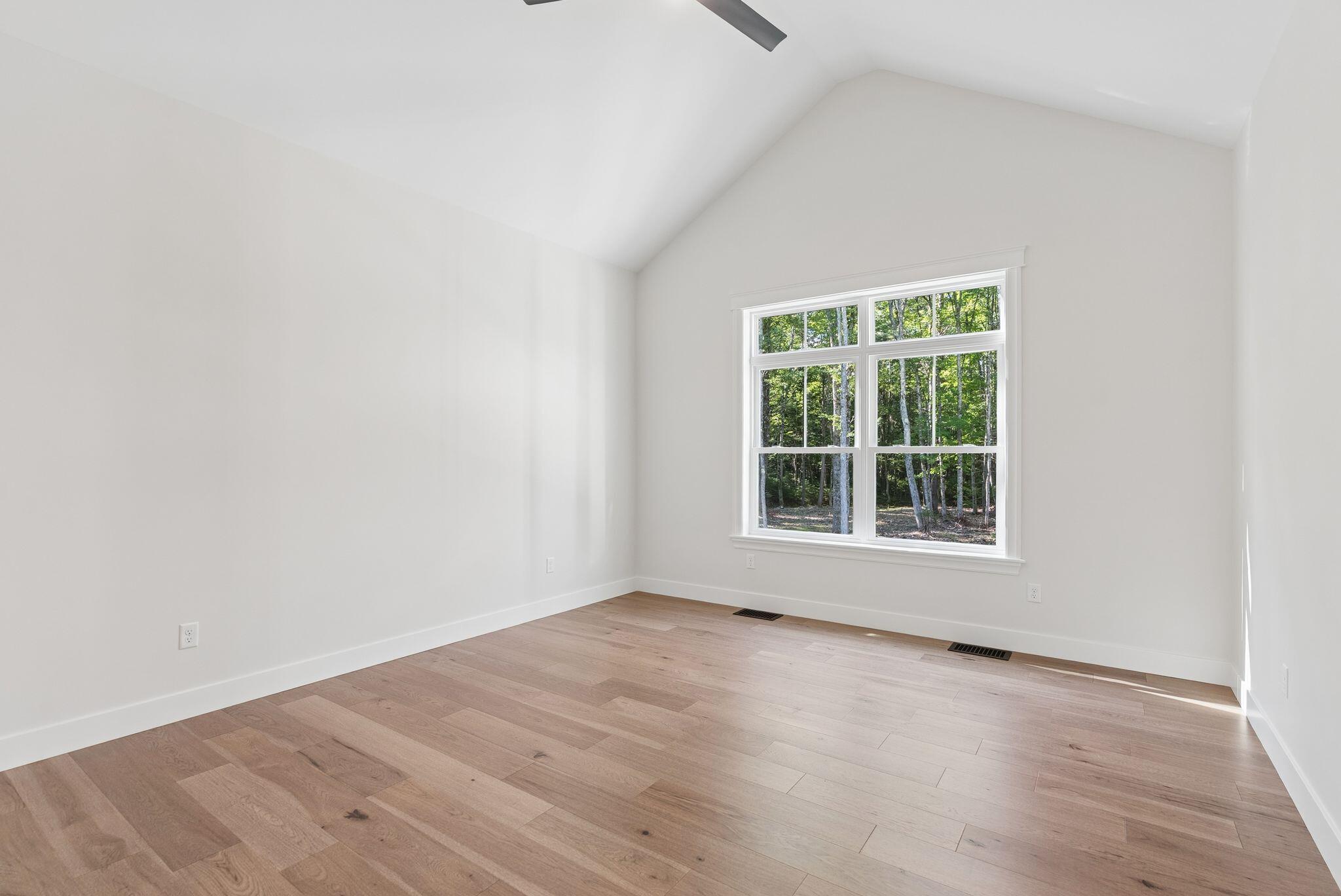 Vacant room with vaulted ceiling, wood floors, and a large window overlooking trees.
