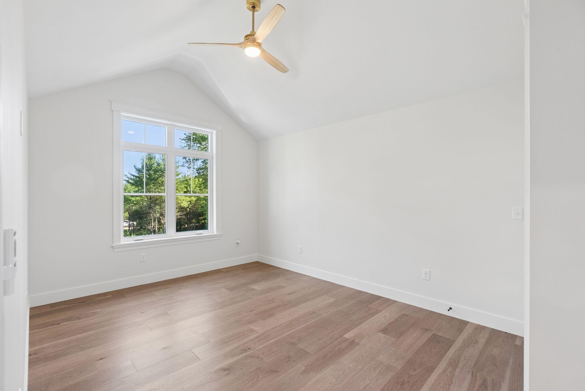 Vacant room with vaulted ceiling, wood floors, a ceiling fan, and a large window overlooking trees.