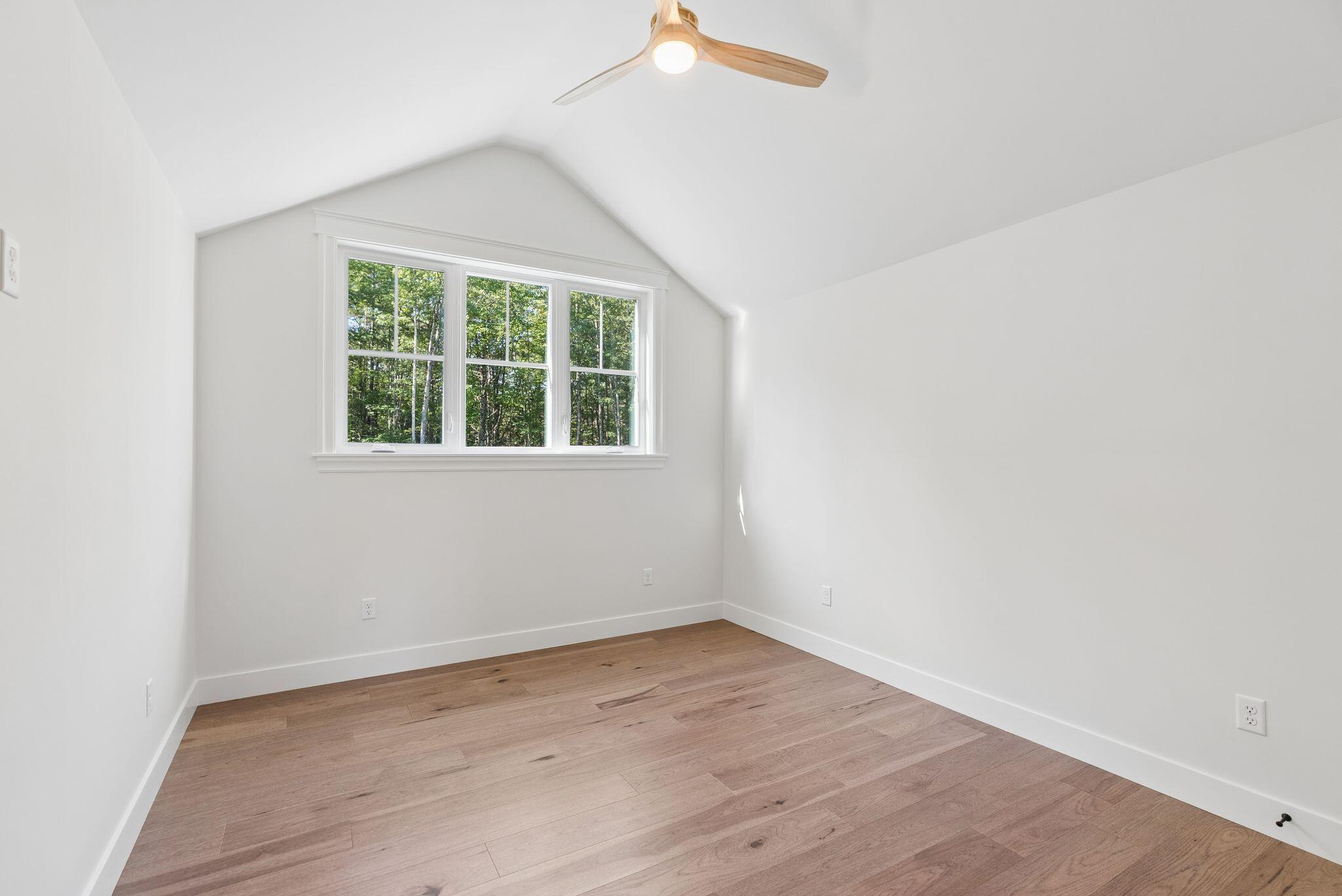 Empty room with wood floors, a vaulted ceiling, and a wide window overlooking trees.