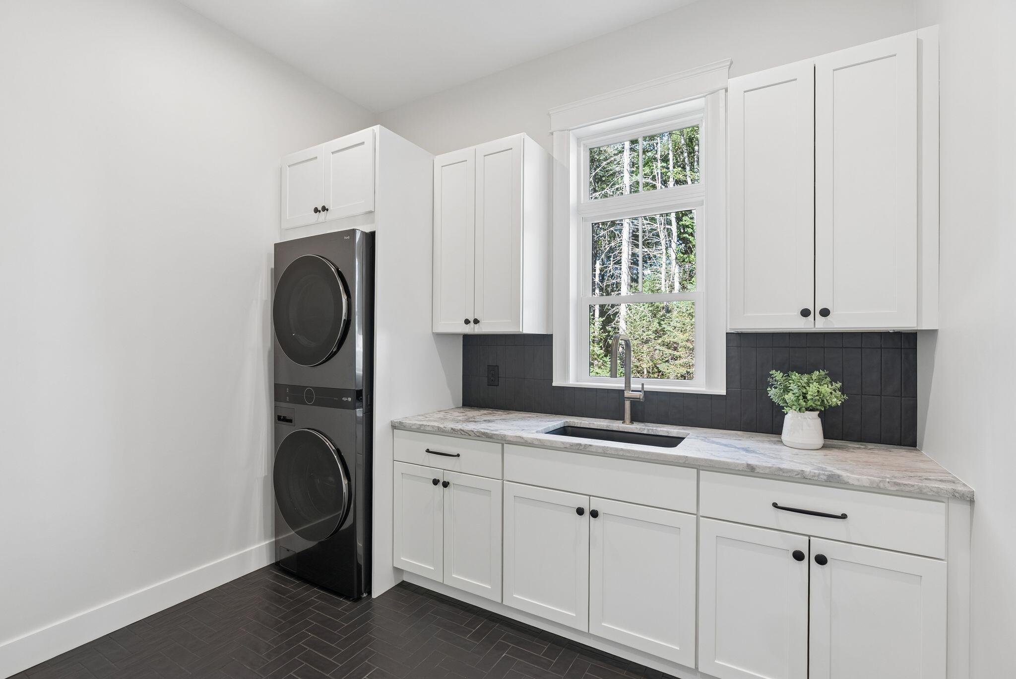 Laundry room with stacked washer and dryer, white cabinetry, and a sink beneath a wooded-view window.