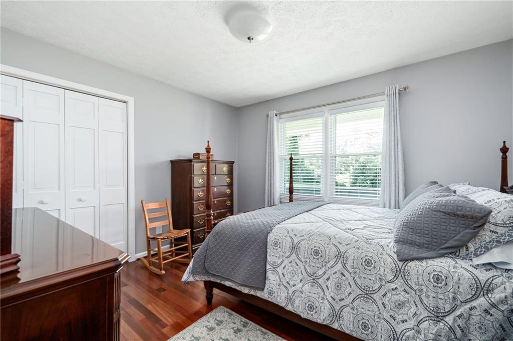 Furnished bedroom with a patterned bed, hardwood floors, and a wide curtained window.