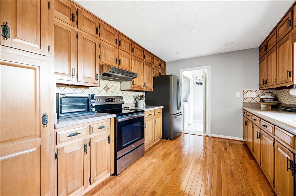 Galley kitchen with honey-toned cabinets, a black range, stainless-steel refrigerator, and hardwood floors.