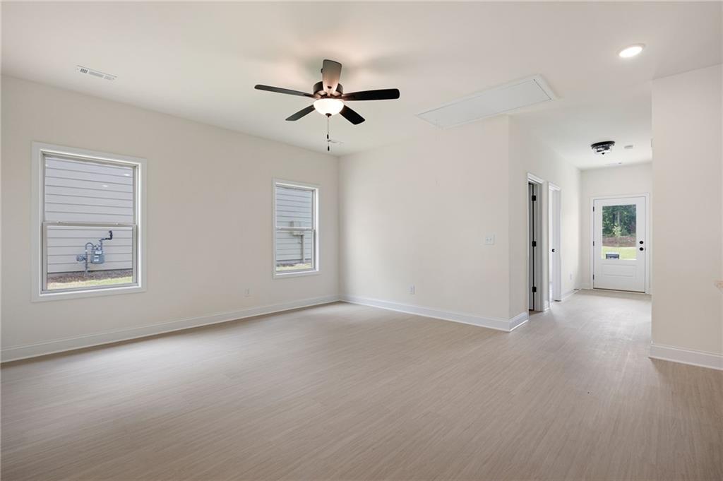 Empty living room with pale flooring, two windows, and a ceiling fan beside a hallway to the exterior door.