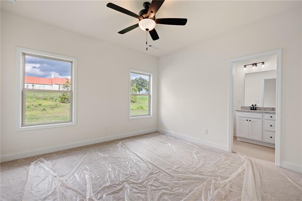 Empty bedroom with carpet covered in plastic, two windows, and a ceiling fan; bathroom visible through doorway.