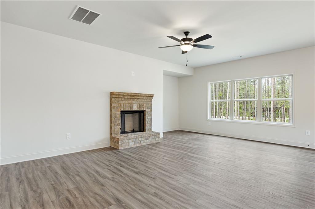 Empty living room with a brick fireplace, gray flooring, ceiling fan, and large window overlooking trees.
