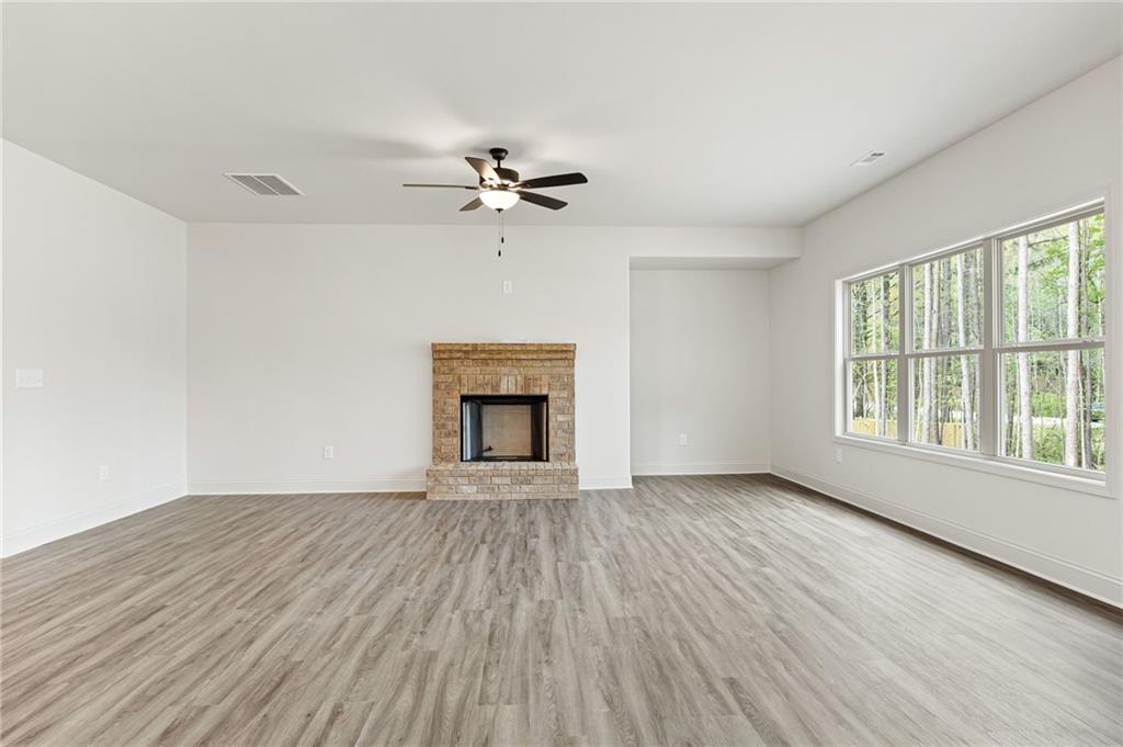 Empty living room with a brick fireplace, gray flooring, and large windows overlooking trees.