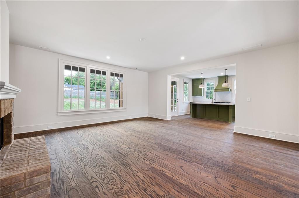 Empty living room with hardwood floors, brick fireplace, and view into a green kitchen.