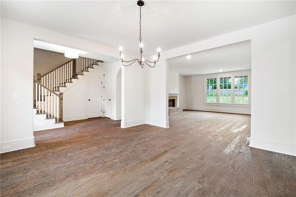 Empty open living and dining area with hardwood floors, staircase, chandelier, and fireplace by large windows.