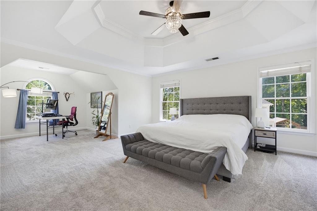 Furnished bedroom with a gray upholstered bed, arched window, and desk nook beneath a tray ceiling.