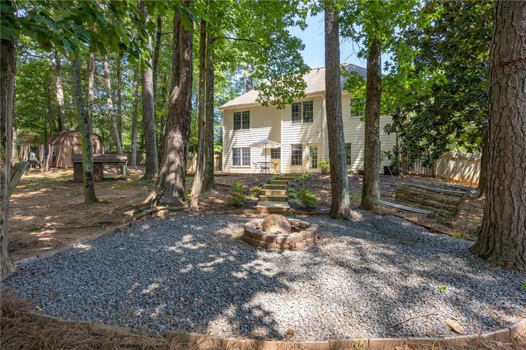 Two-story home beyond a wooded backyard with a gravel fire pit area and wooden bench.
