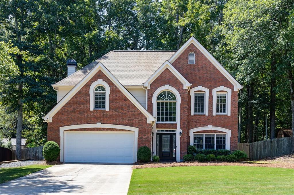 Two-story brick house with a double garage, white trim, and mature trees behind it.