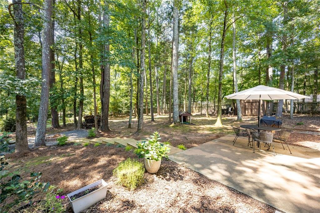 Wooded backyard patio with umbrella-shaded dining set, mature trees, and a fenced yard.