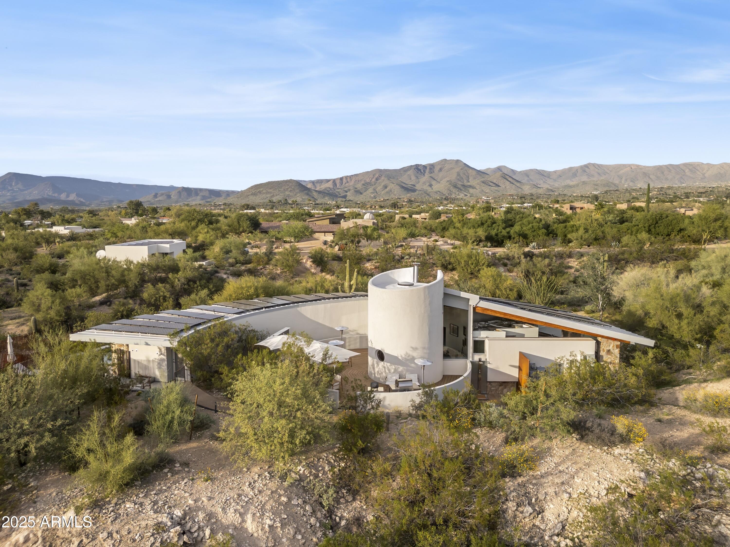 Modern desert home with a cylindrical tower and rooftop panels, surrounded by desert vegetation beneath a mountain range.