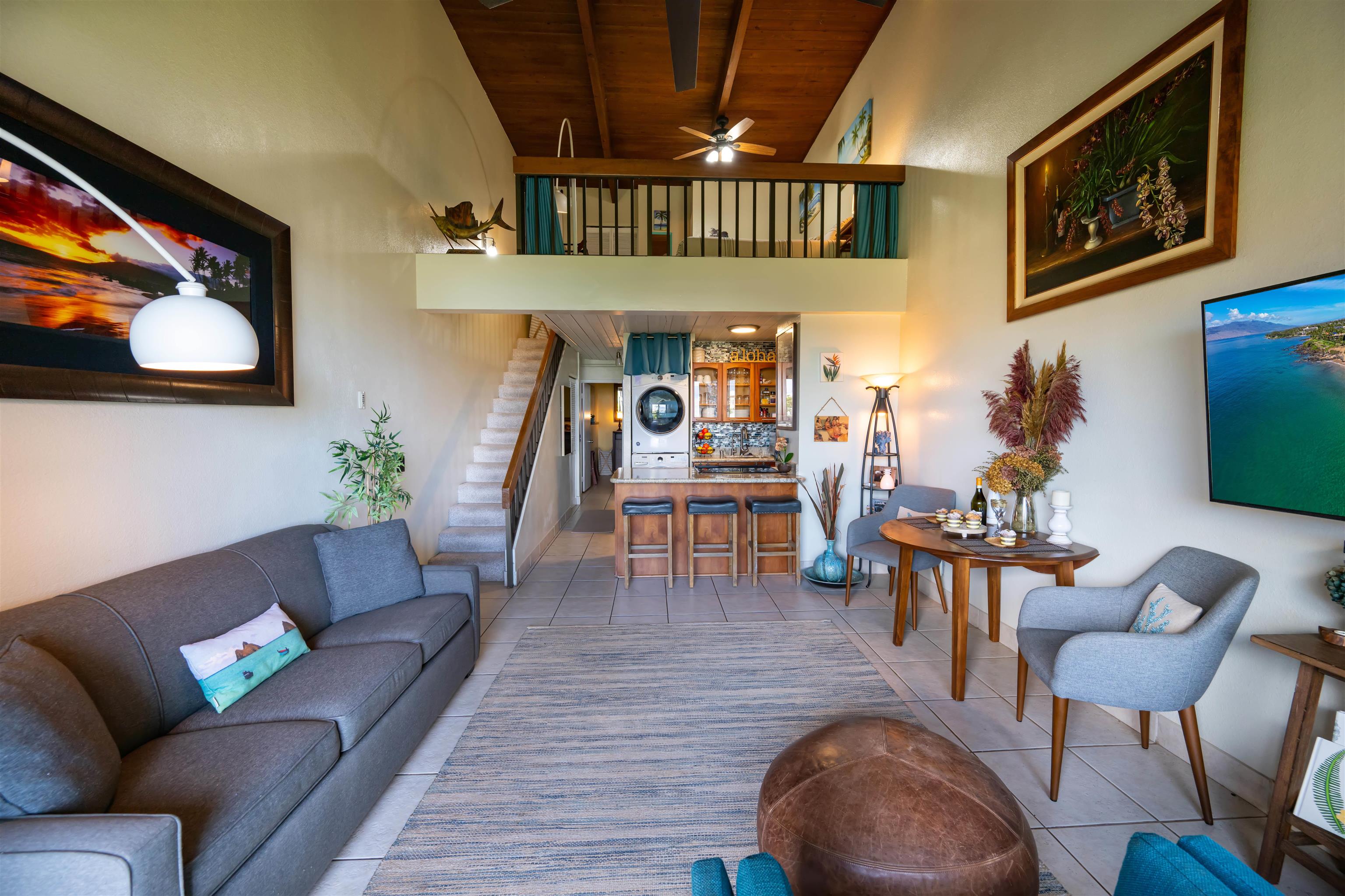 Furnished condo living room with a gray sofa, open kitchen, staircase, and loft beneath a wood ceiling.
