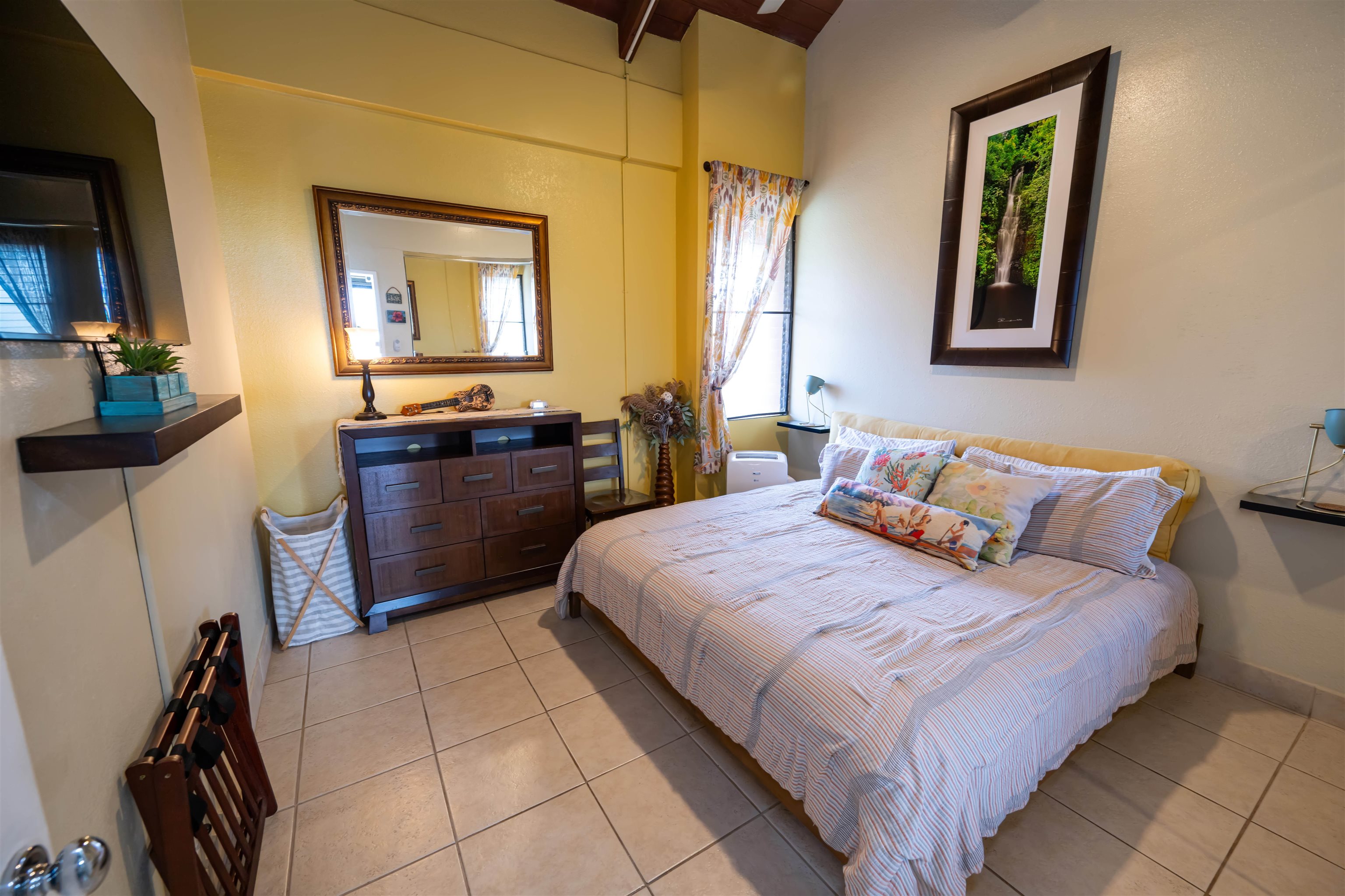 Furnished bedroom with a striped bed, colorful pillows, dark wood dresser, and tiled floor.