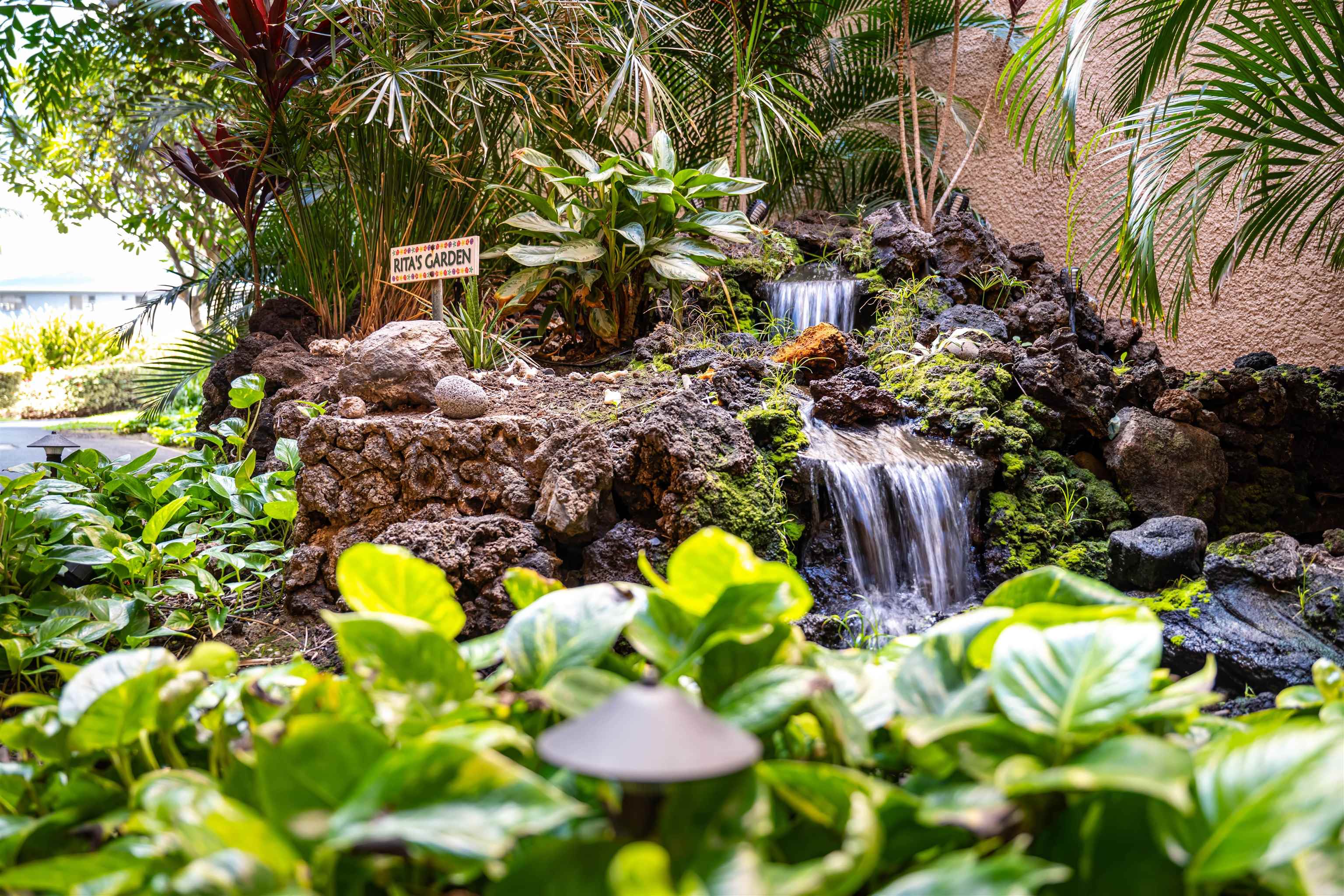 Tropical garden waterfall surrounded by rocks and lush plants, with a “Rita’s Garden” sign.