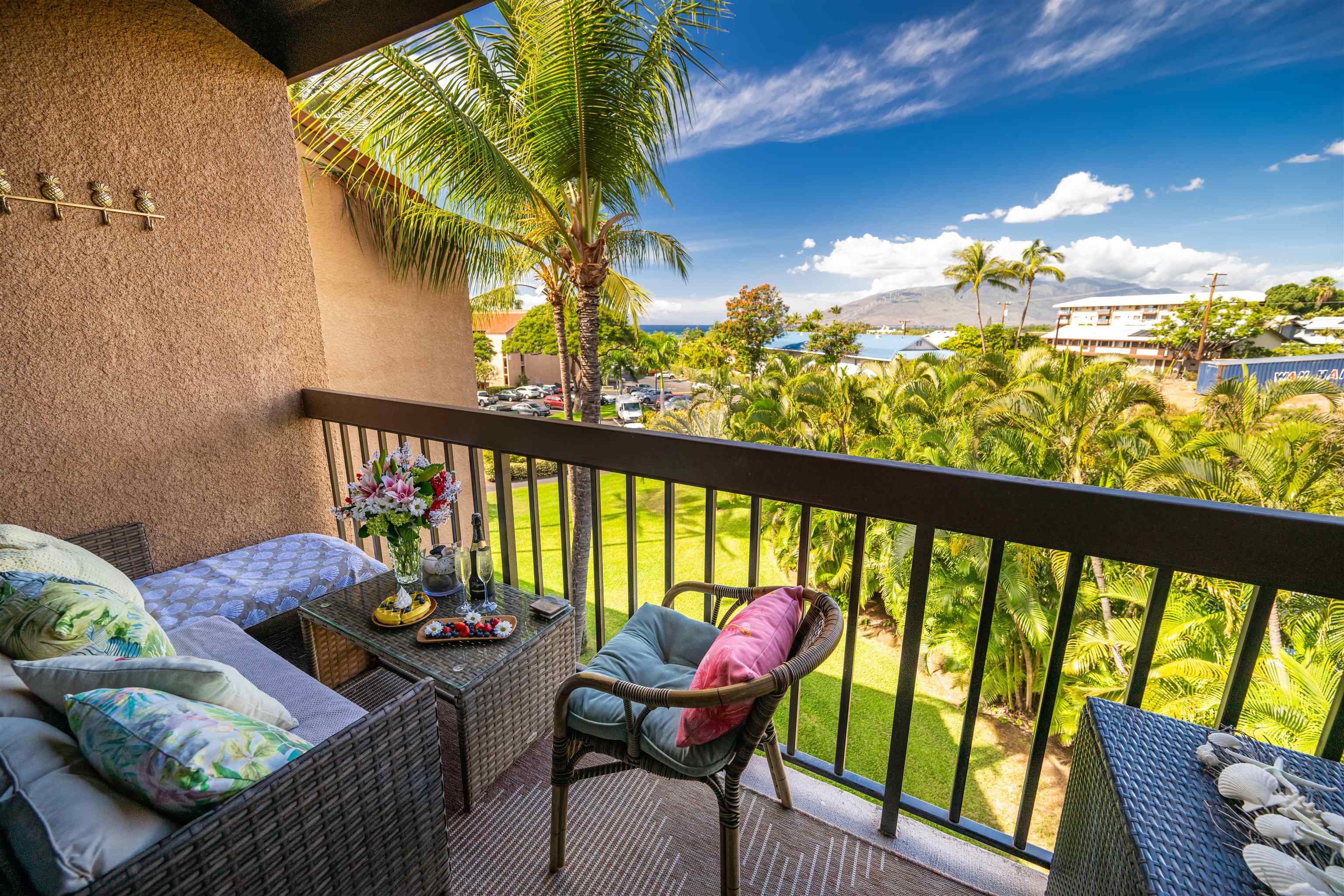 Furnished balcony overlooks palm trees, neighboring buildings, ocean, and a distant mountain.