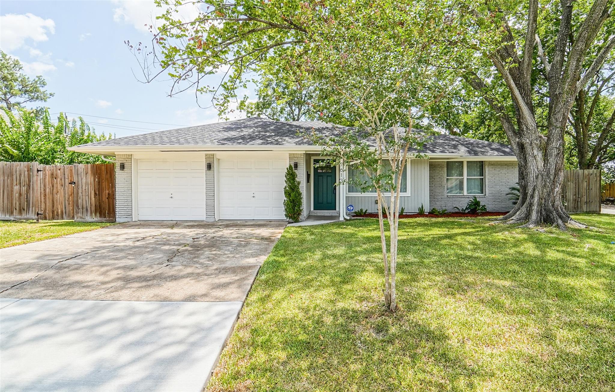 Single-story brick house with a two-car garage, green front door, broad driveway, and mature shade tree.