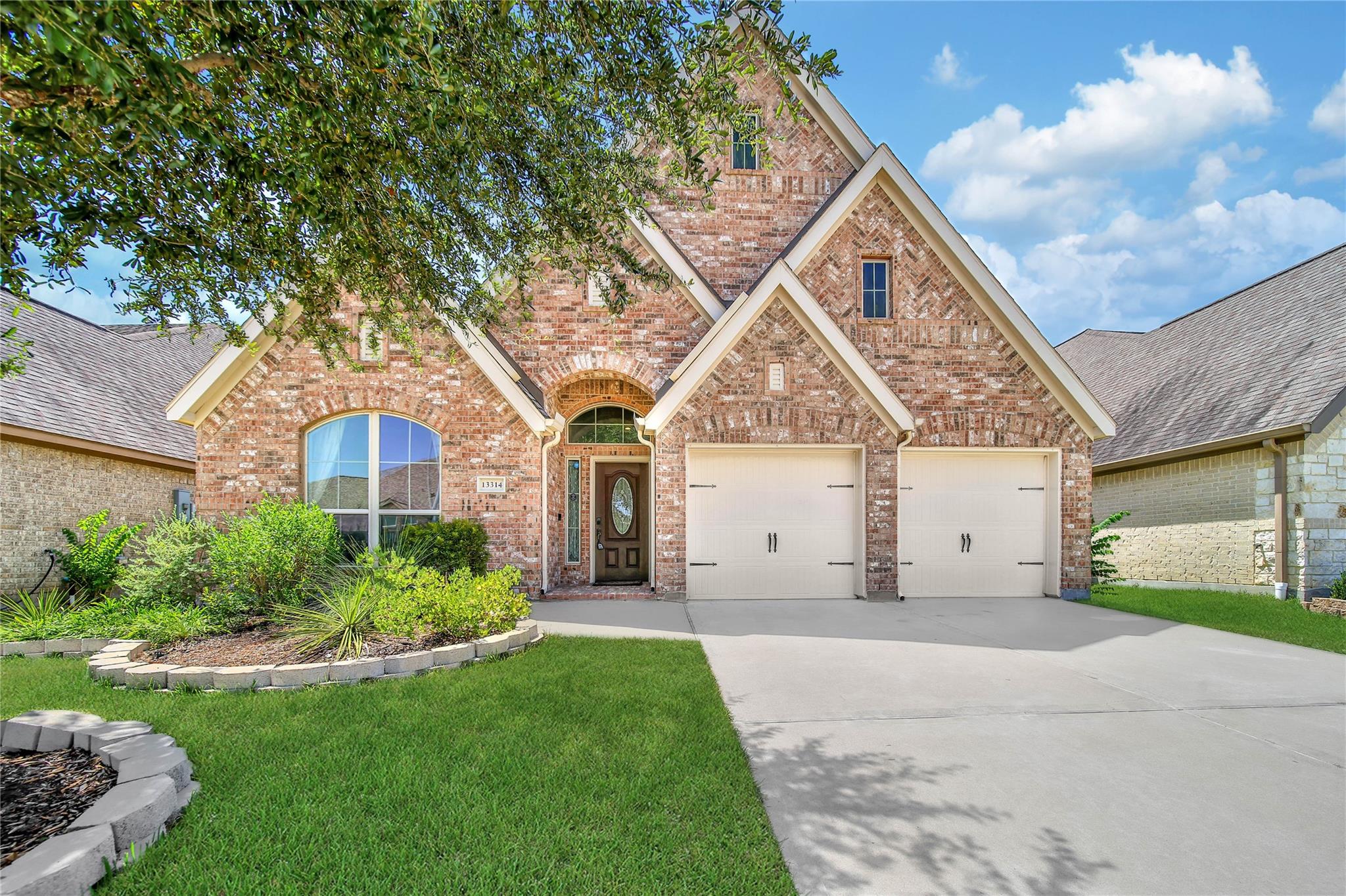 Red brick home with steep gables, two garage doors, landscaped lawn, and concrete driveway.