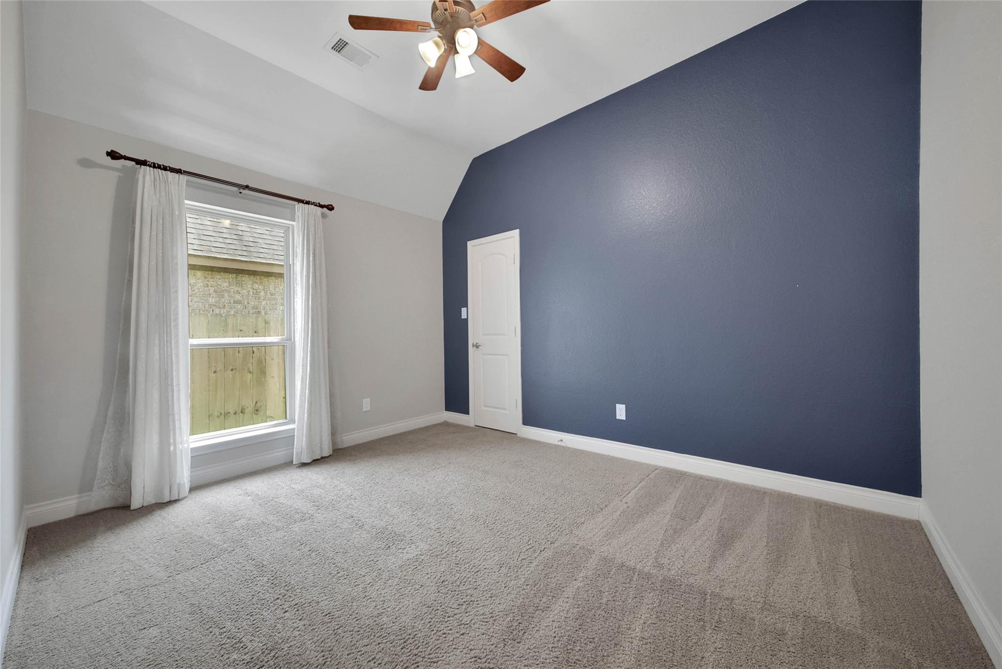 Empty bedroom with a dark blue accent wall, beige carpet, and curtained window.