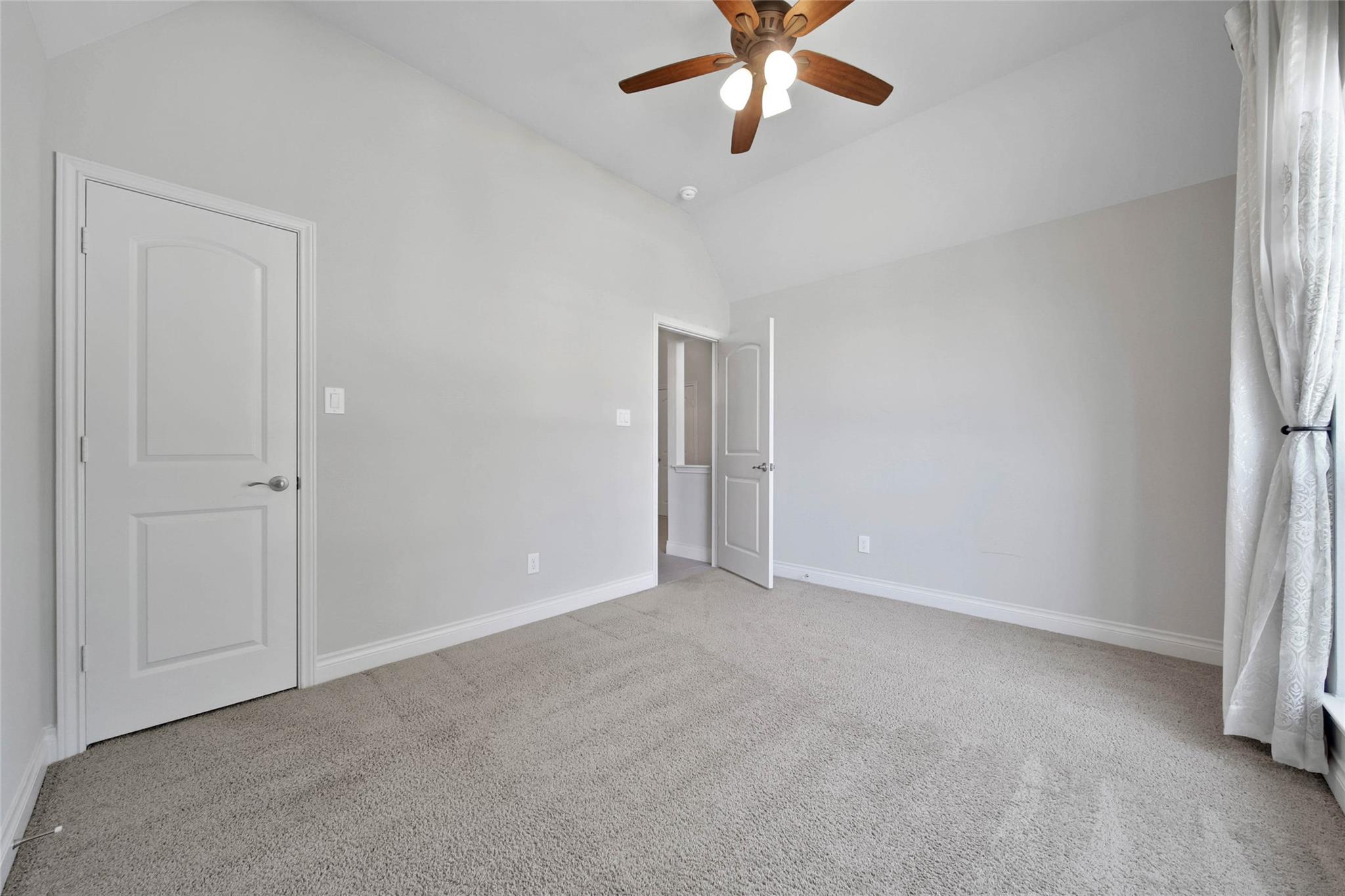 Empty carpeted bedroom with a wooden-blade ceiling fan and white curtains.