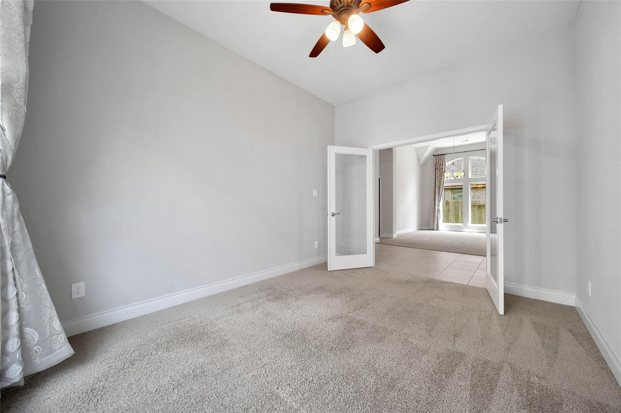 Empty carpeted bedroom with a ceiling fan and open double doors to a bright adjoining room.