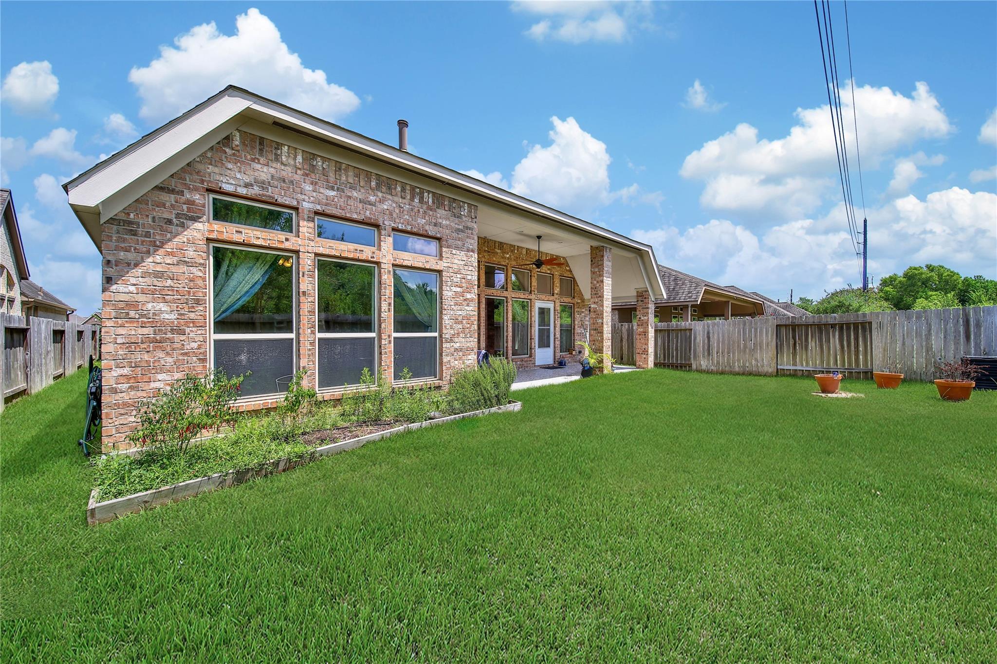Brick house rear exterior with covered patio and tall windows overlooking a fenced, grassy backyard.