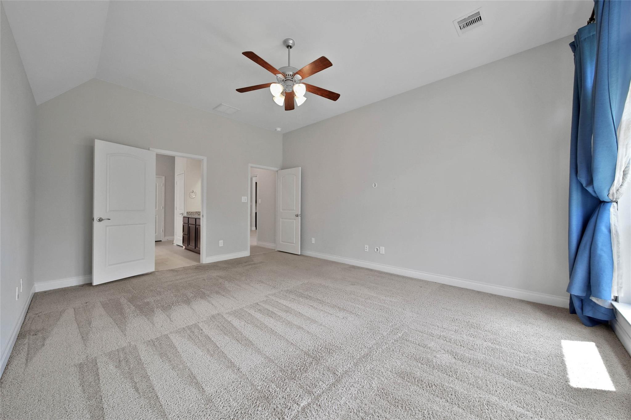 Empty carpeted bedroom with a ceiling fan, blue curtains, and open doors to adjoining rooms.