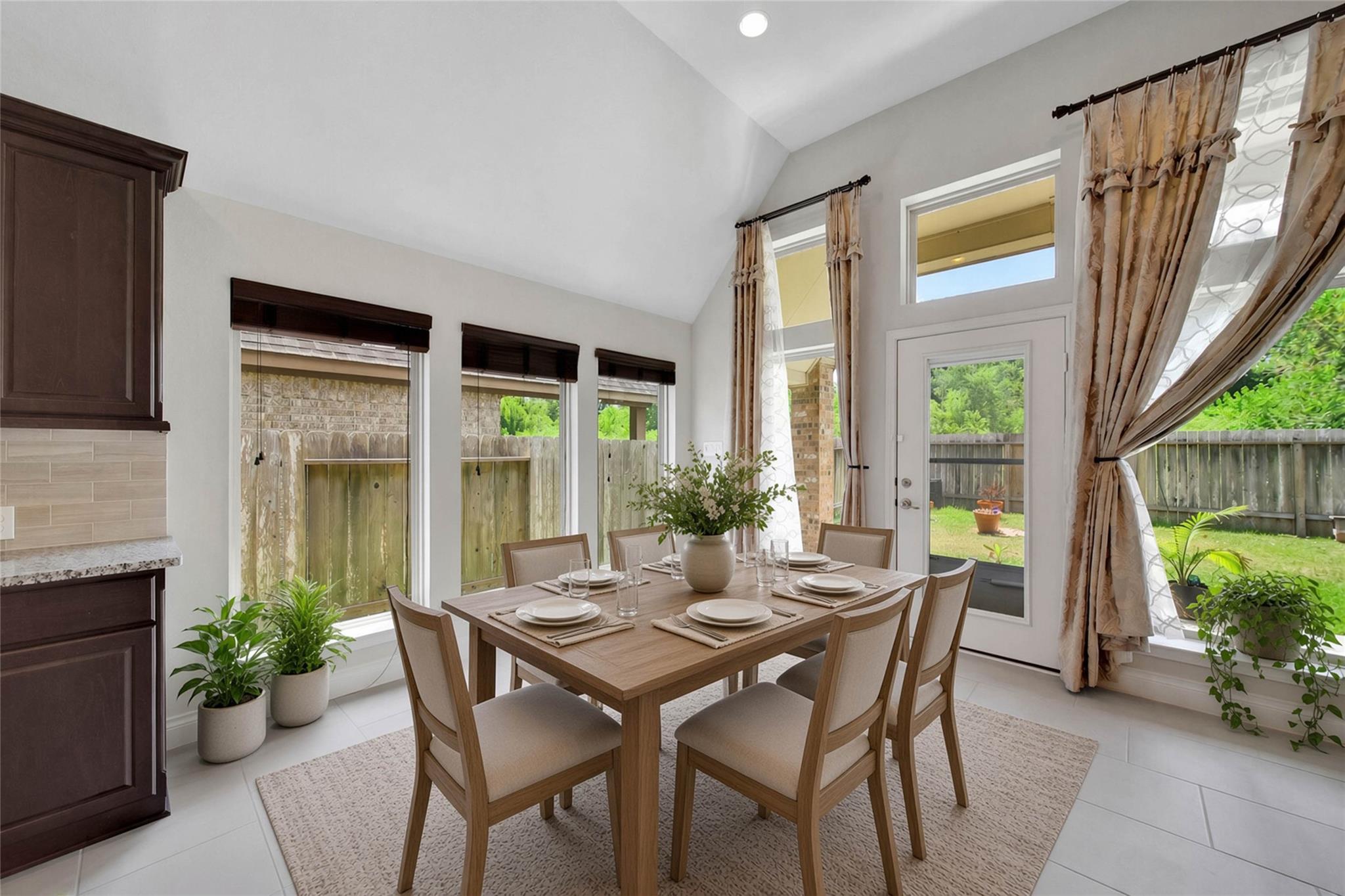 Dining area with a set wooden table beneath a vaulted ceiling, beside large windows and a glass backyard door.
