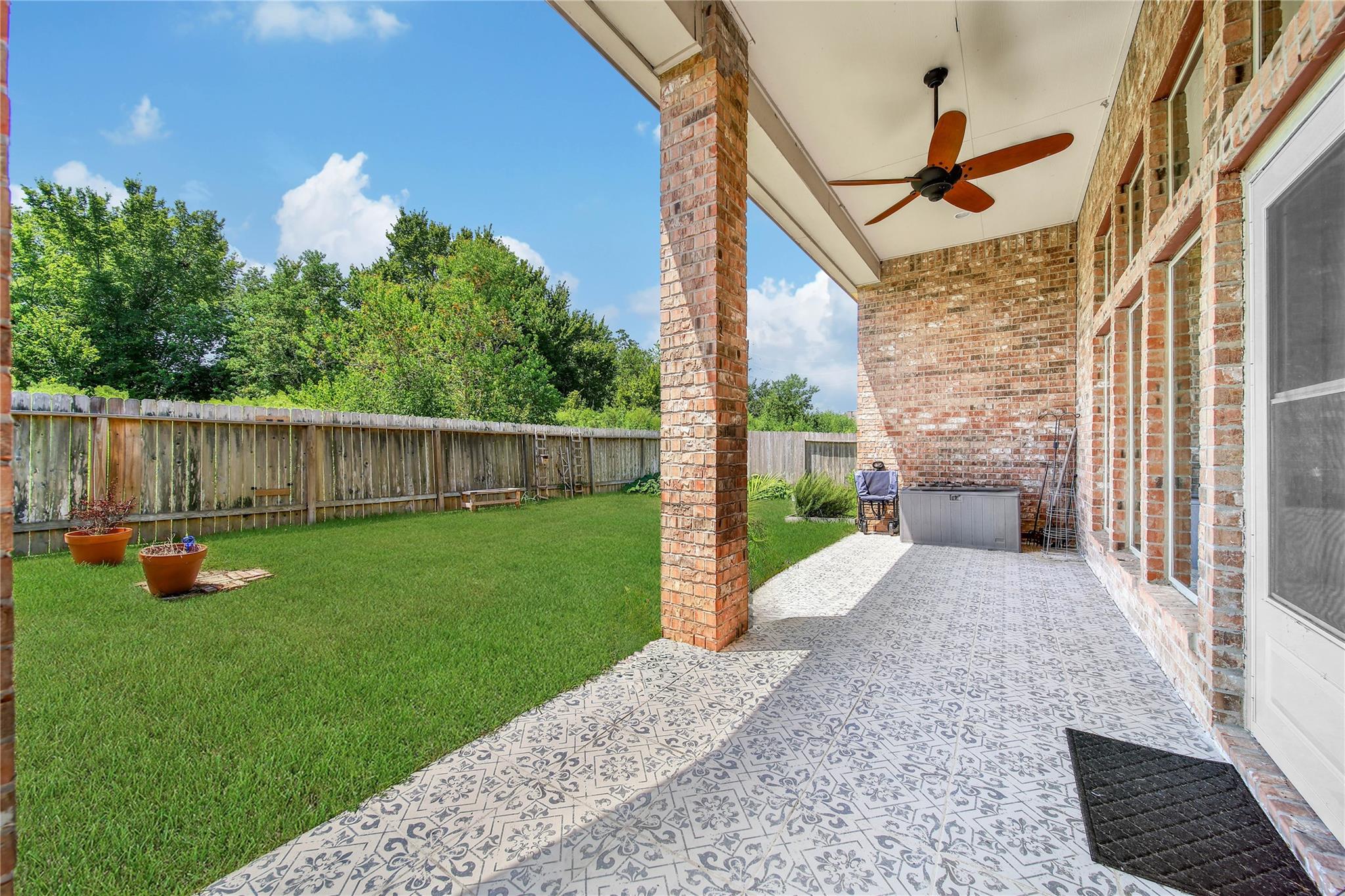 Covered brick patio with patterned tile, ceiling fan, and a view of the fenced lawn.