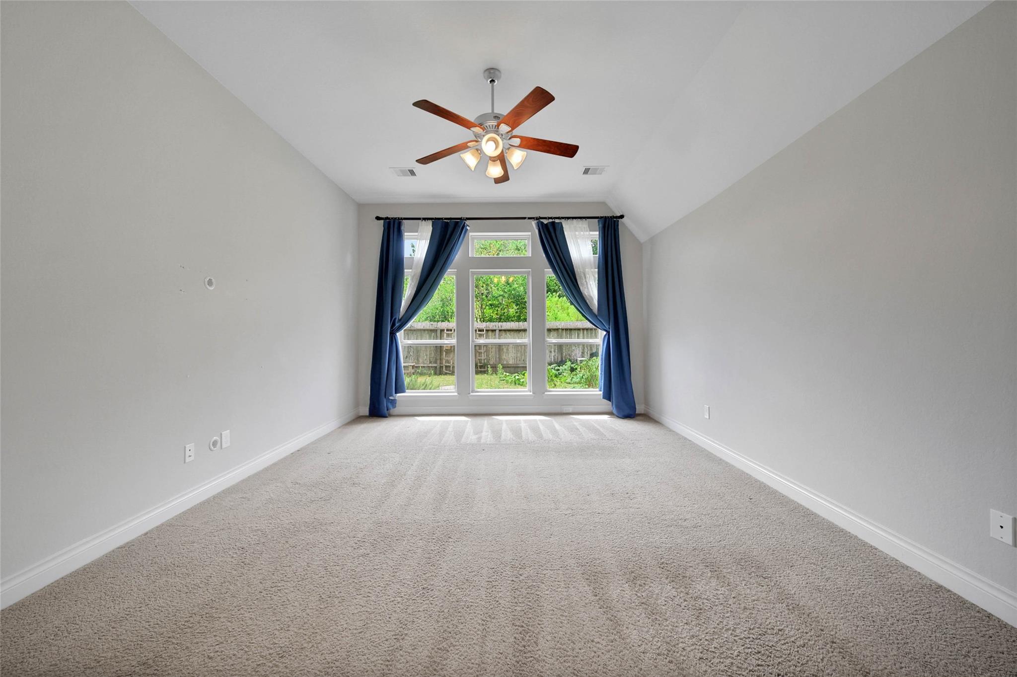 Empty carpeted room with a vaulted ceiling, ceiling fan, and tall windows with blue curtains.