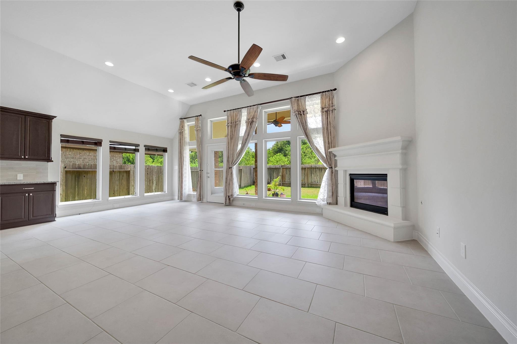 Vacant living room with vaulted ceiling, large backyard-facing windows, tiled floors, and a white fireplace.