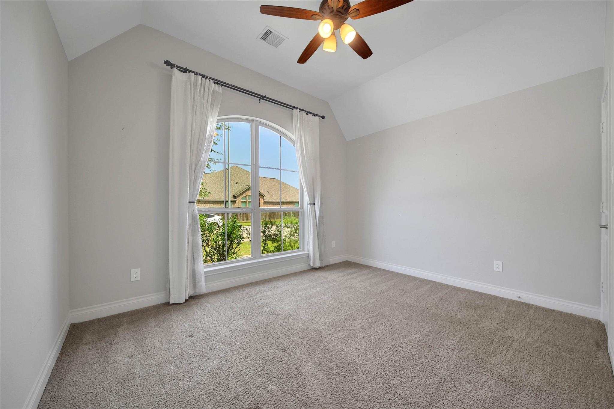 Empty carpeted bedroom with vaulted ceiling, ceiling fan, and large arched window with white curtains.