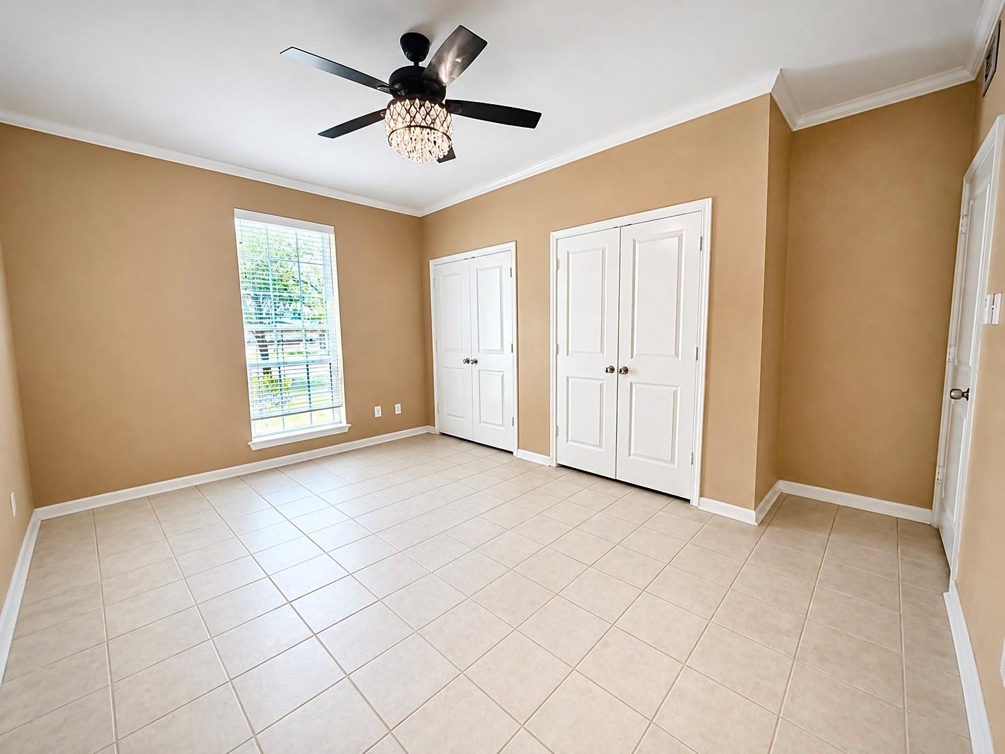 Empty bedroom with tan walls, tiled floor, white closets, and a dark ceiling fan with a crystal-style light.