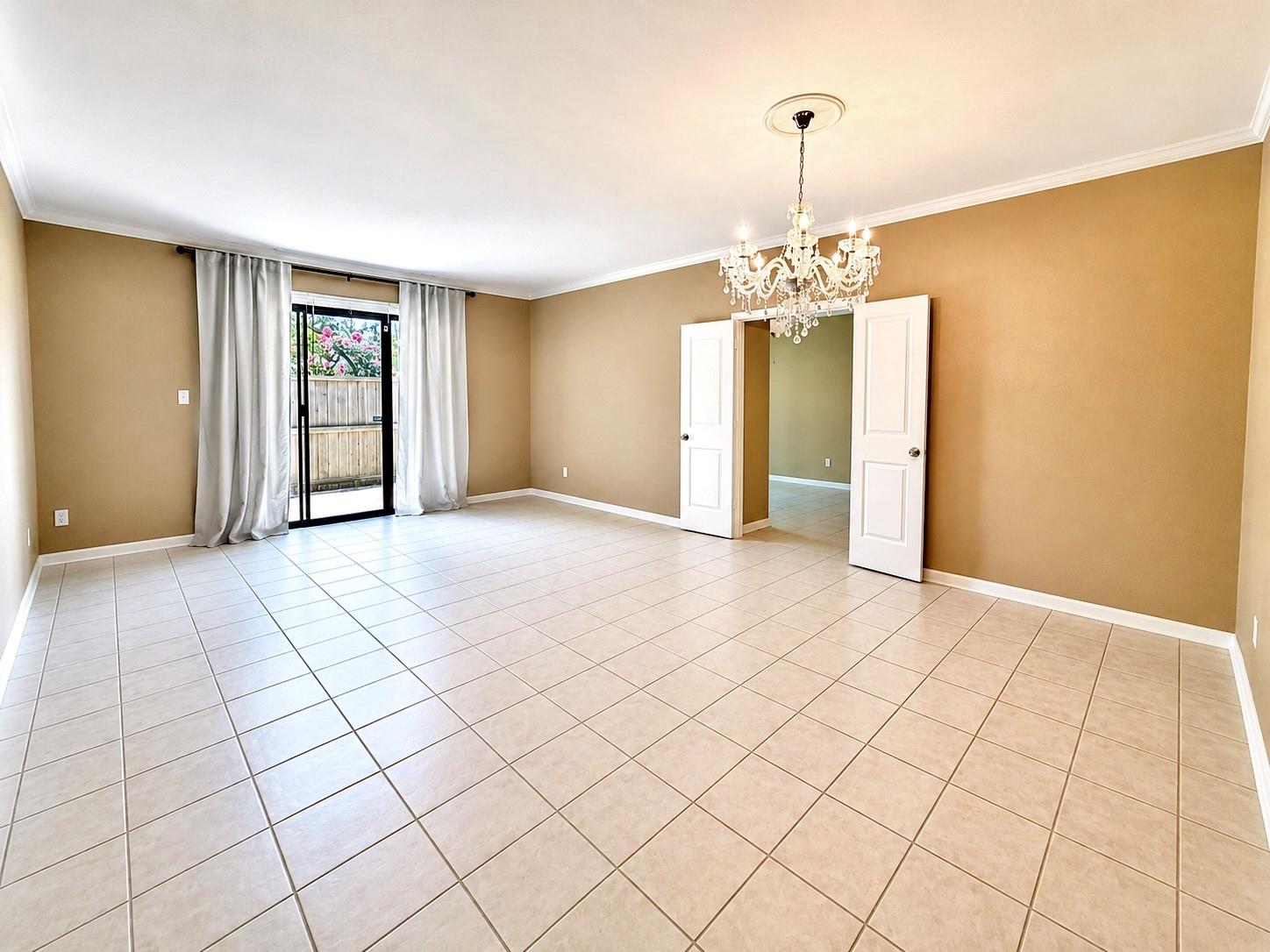 Empty living room with tan walls, tiled floor, chandelier, and sliding glass door to a patio.