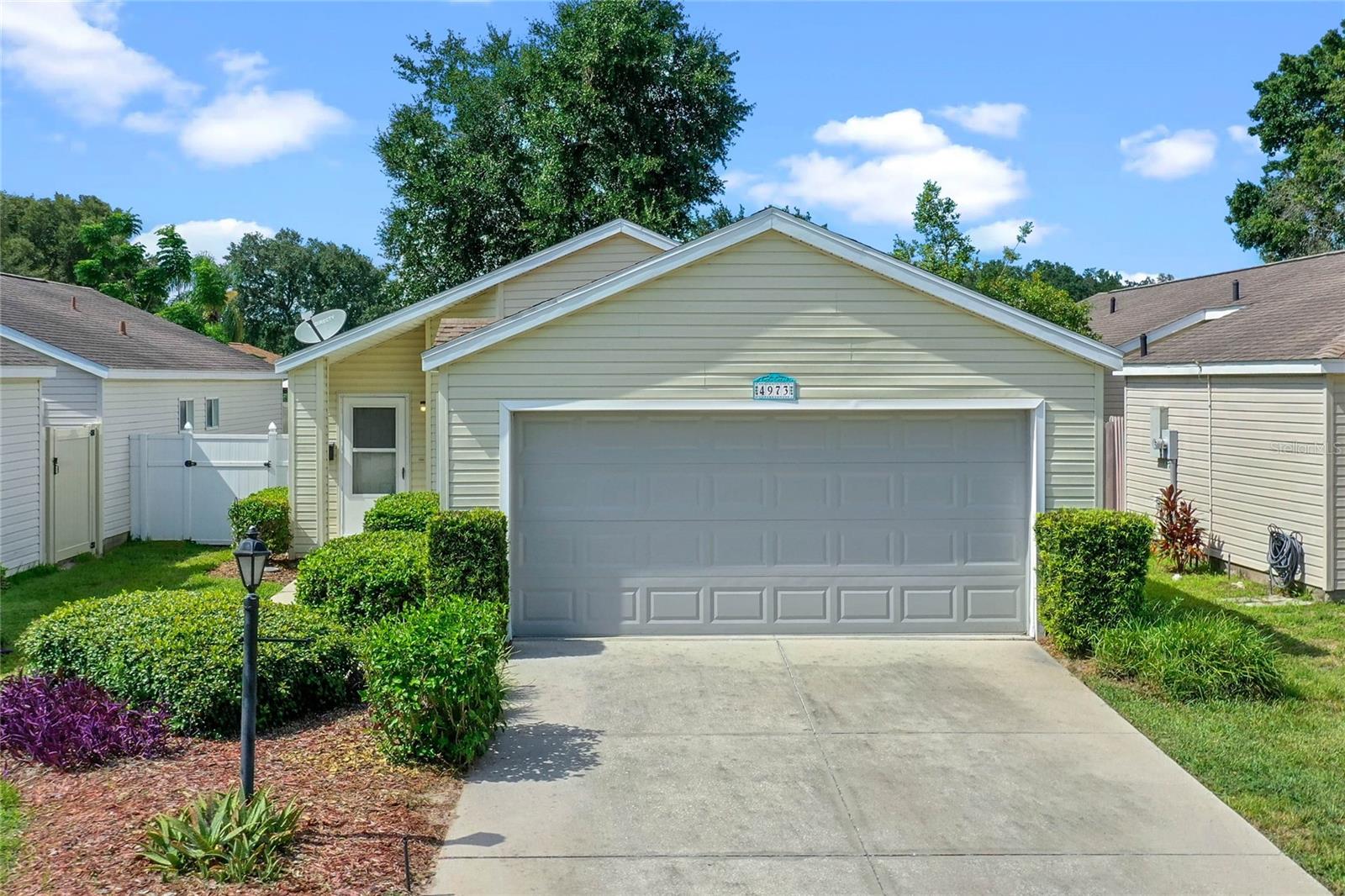 Single-story light-colored house with a two-car garage, landscaped front yard, and driveway.