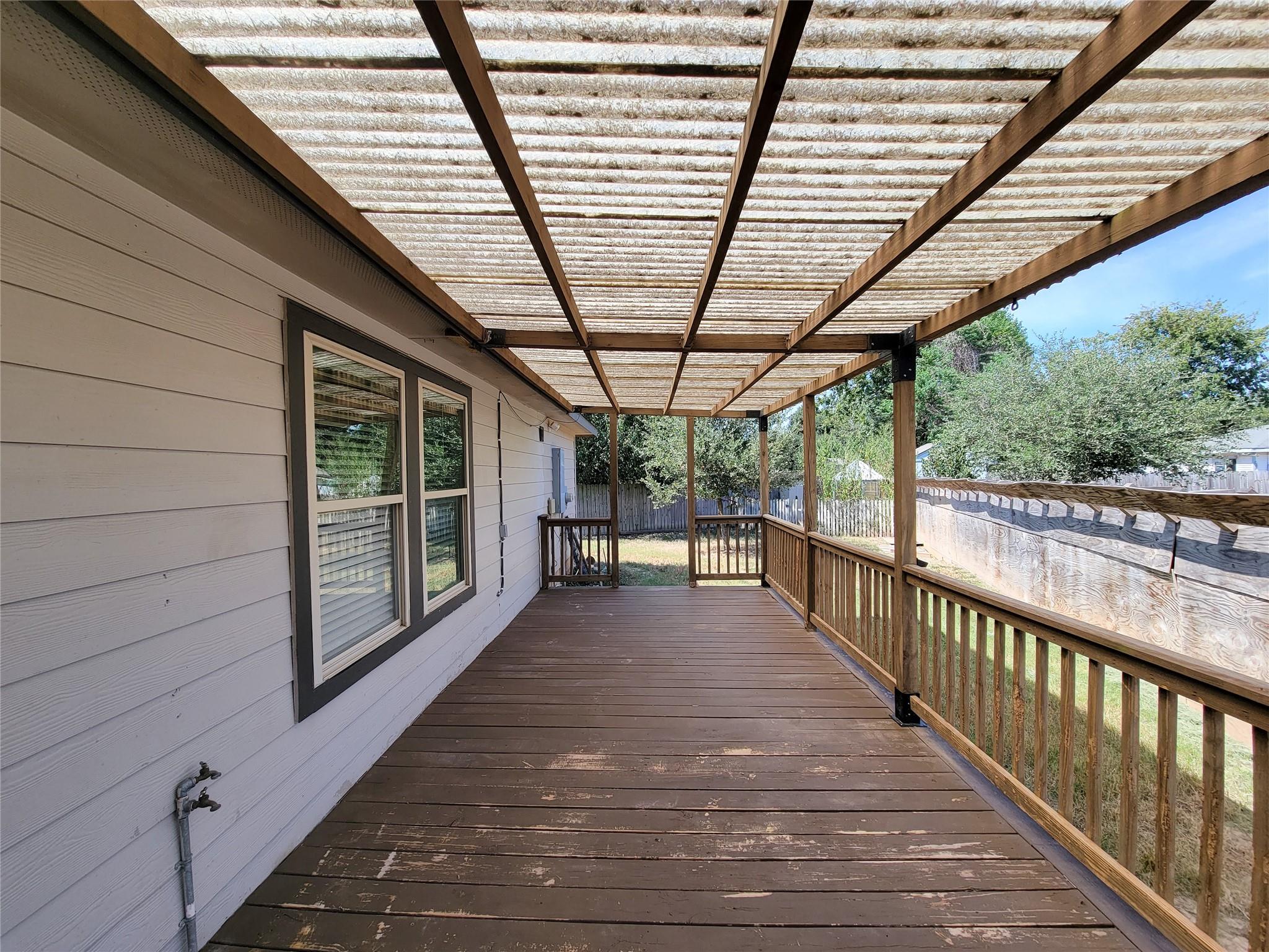 Covered wooden back porch with a translucent pergola roof and railings overlooking a fenced yard.