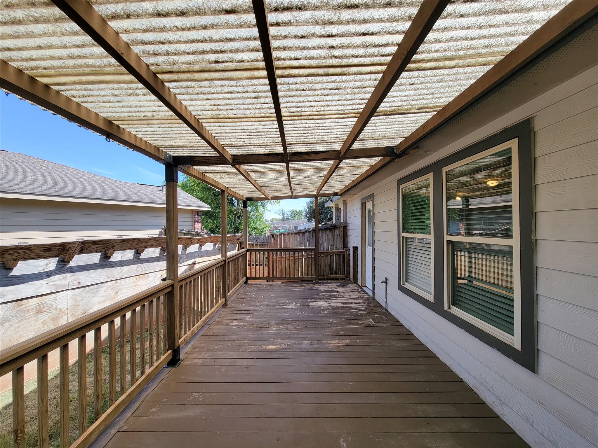 Covered wooden back deck beside the house, with a translucent roof, railings, and large windows.