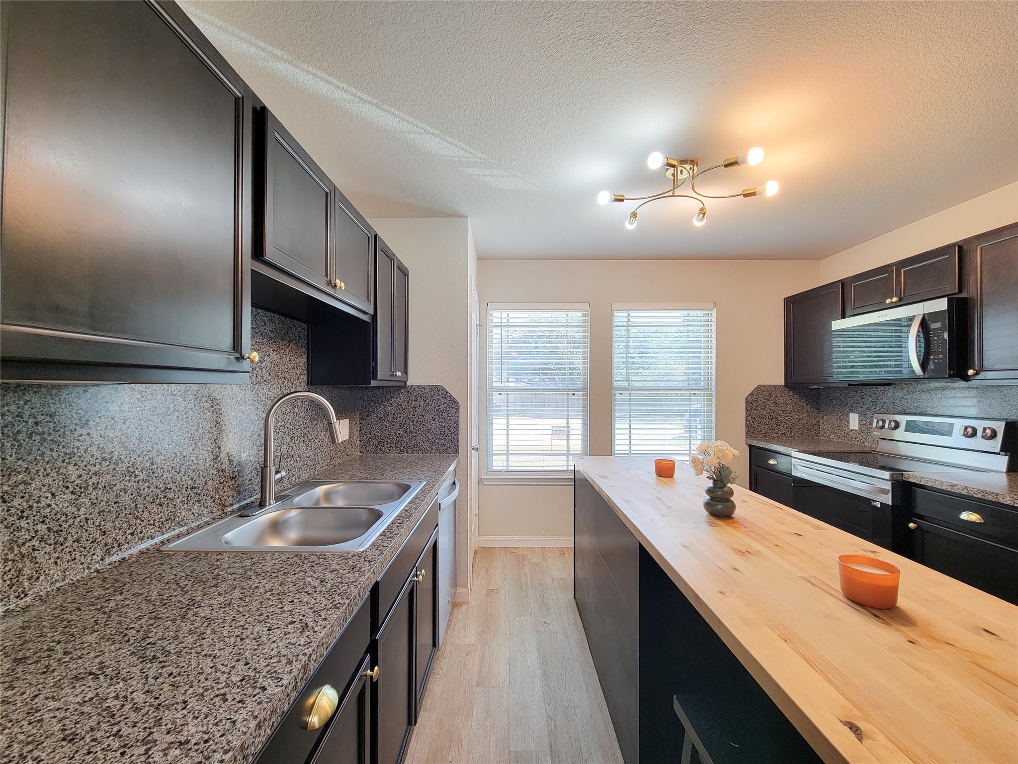 Kitchen with dark cabinets, speckled countertops, stainless appliances, and a long light-wood island.