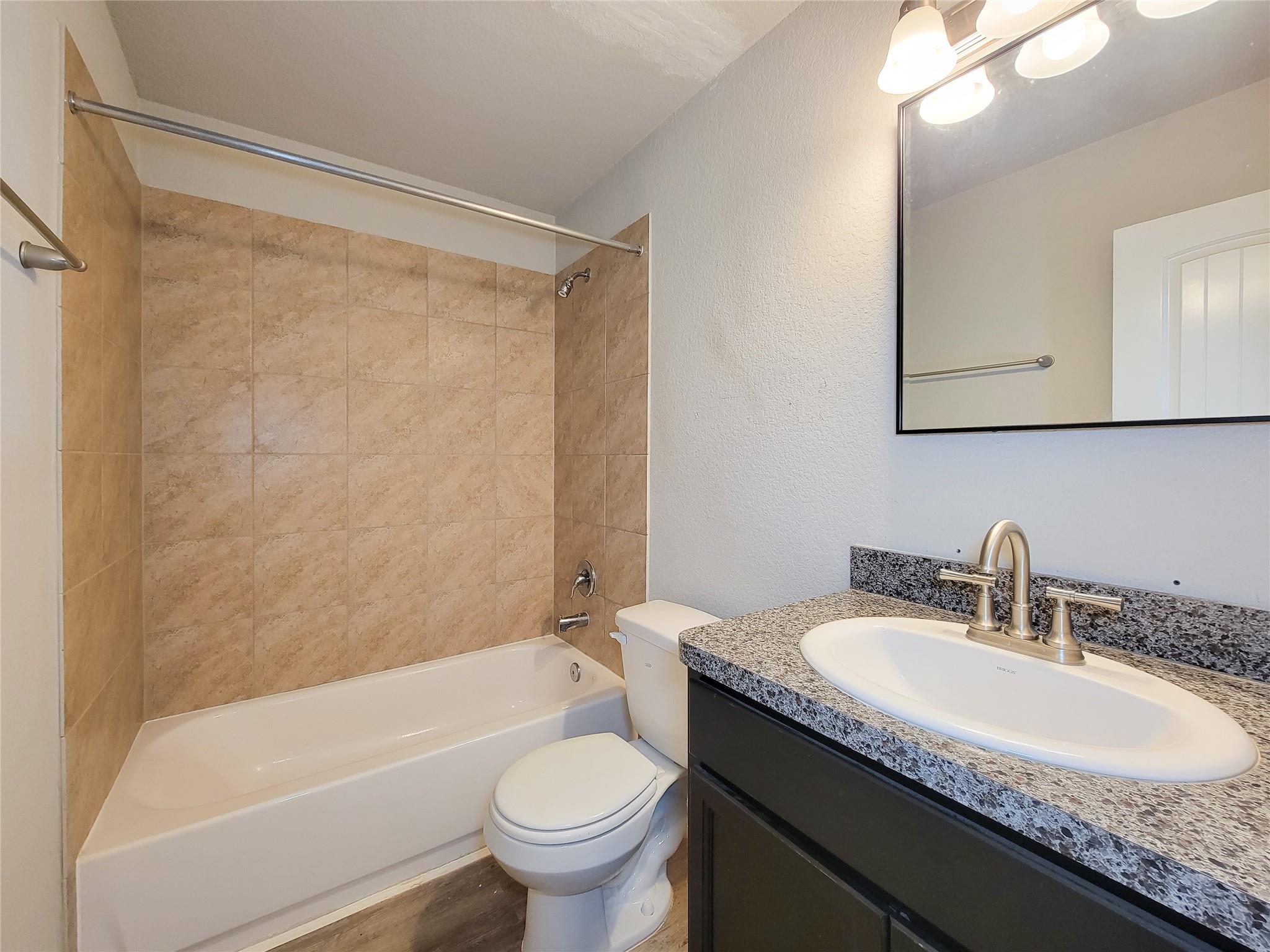 Bathroom with a tan tiled tub and shower beside a dark vanity with granite counter.