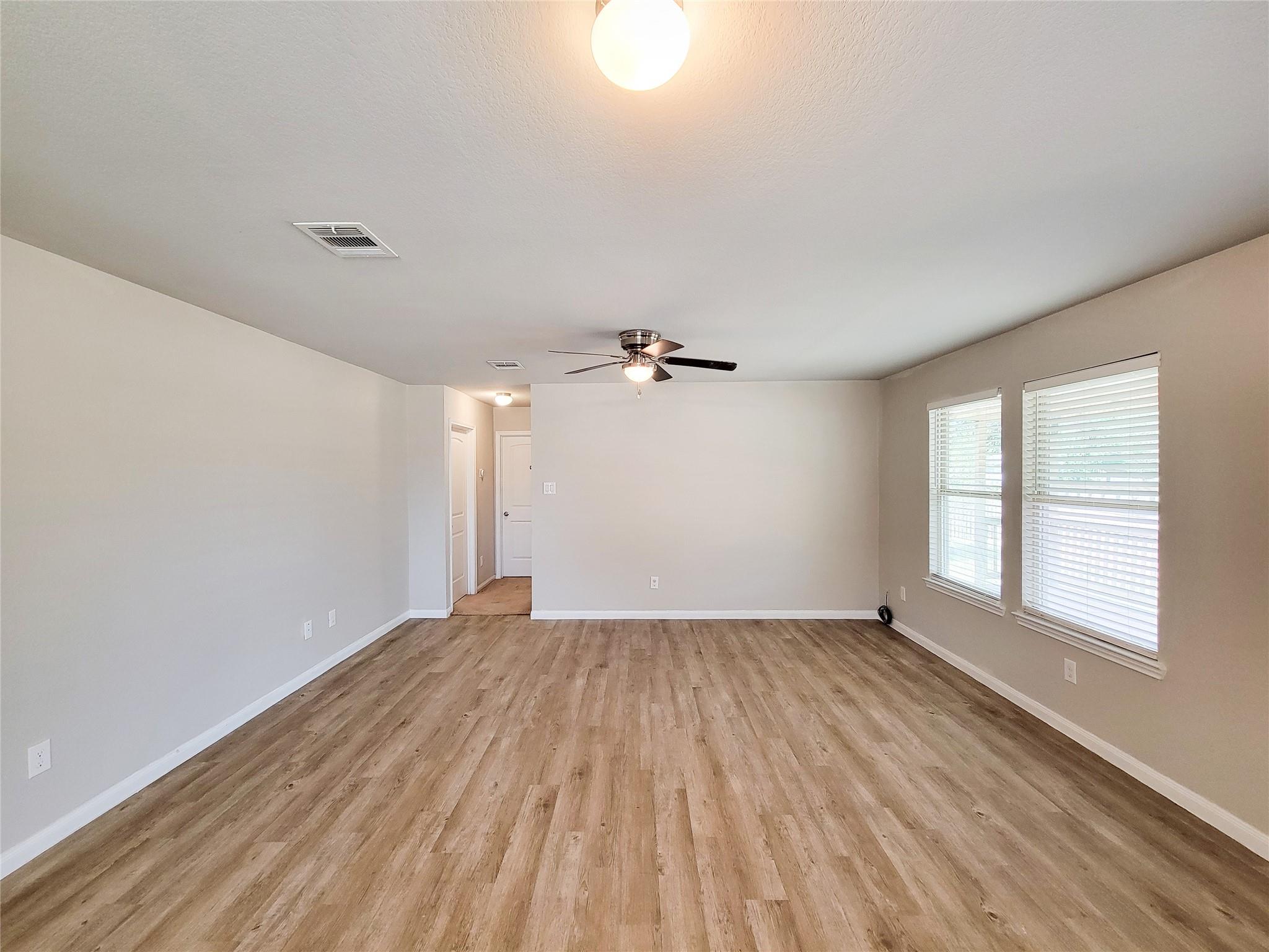 Empty living room with wood-look floors, two windows with blinds, and a ceiling fan.