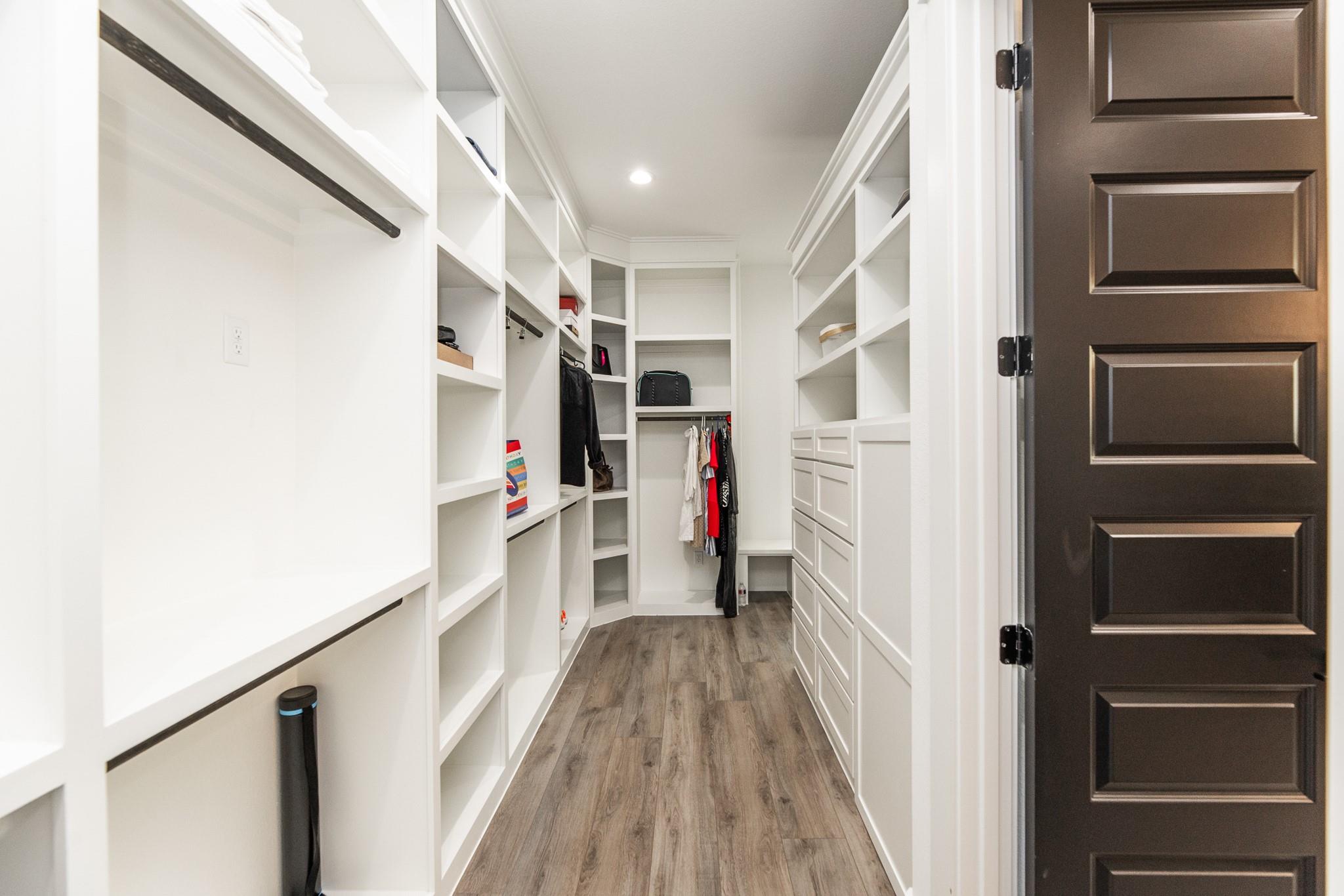 Walk-in closet with white built-in shelves and drawers and wood-look flooring.