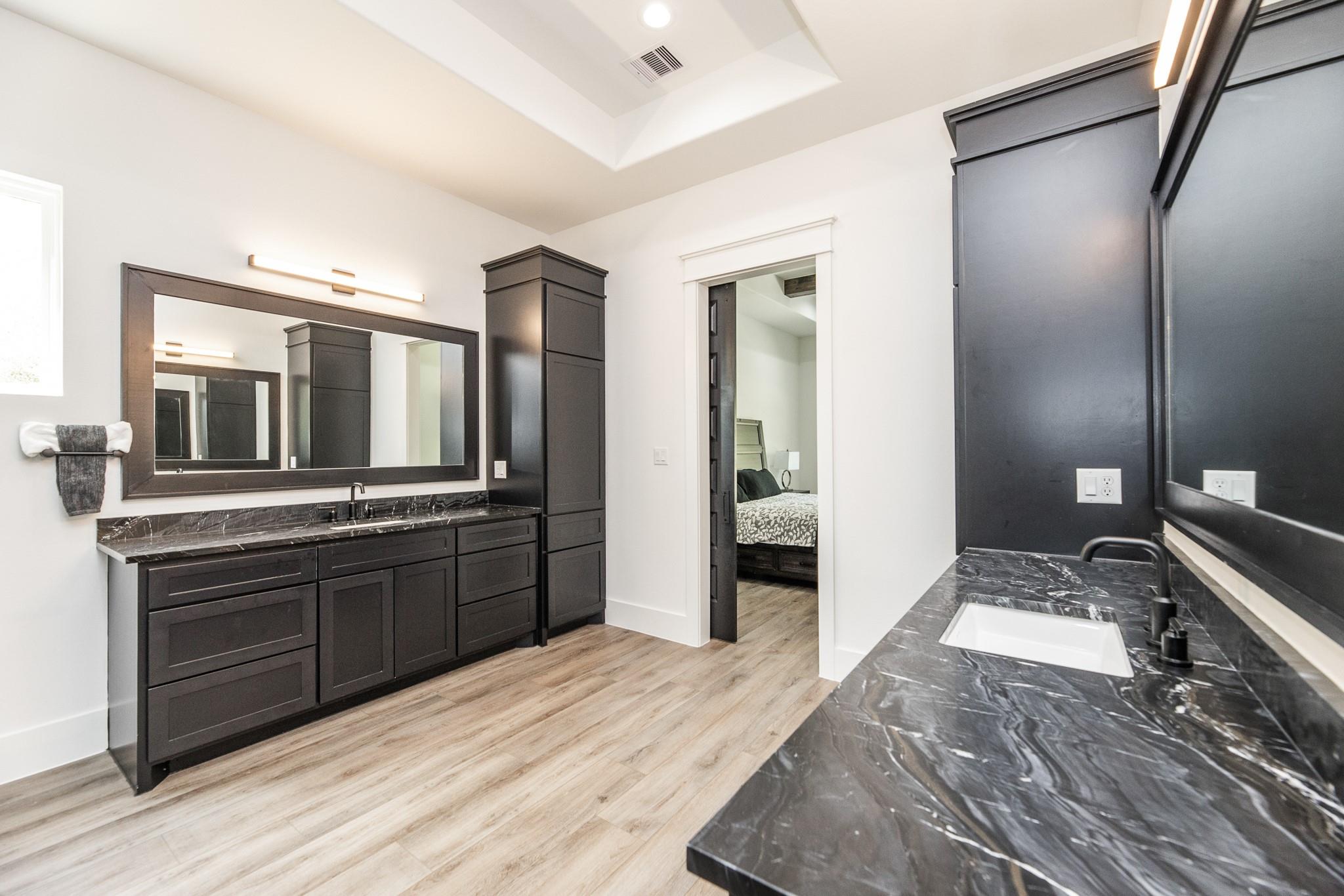 Bathroom with dual dark vanities, marble-look counters, and a view into the adjoining bedroom.