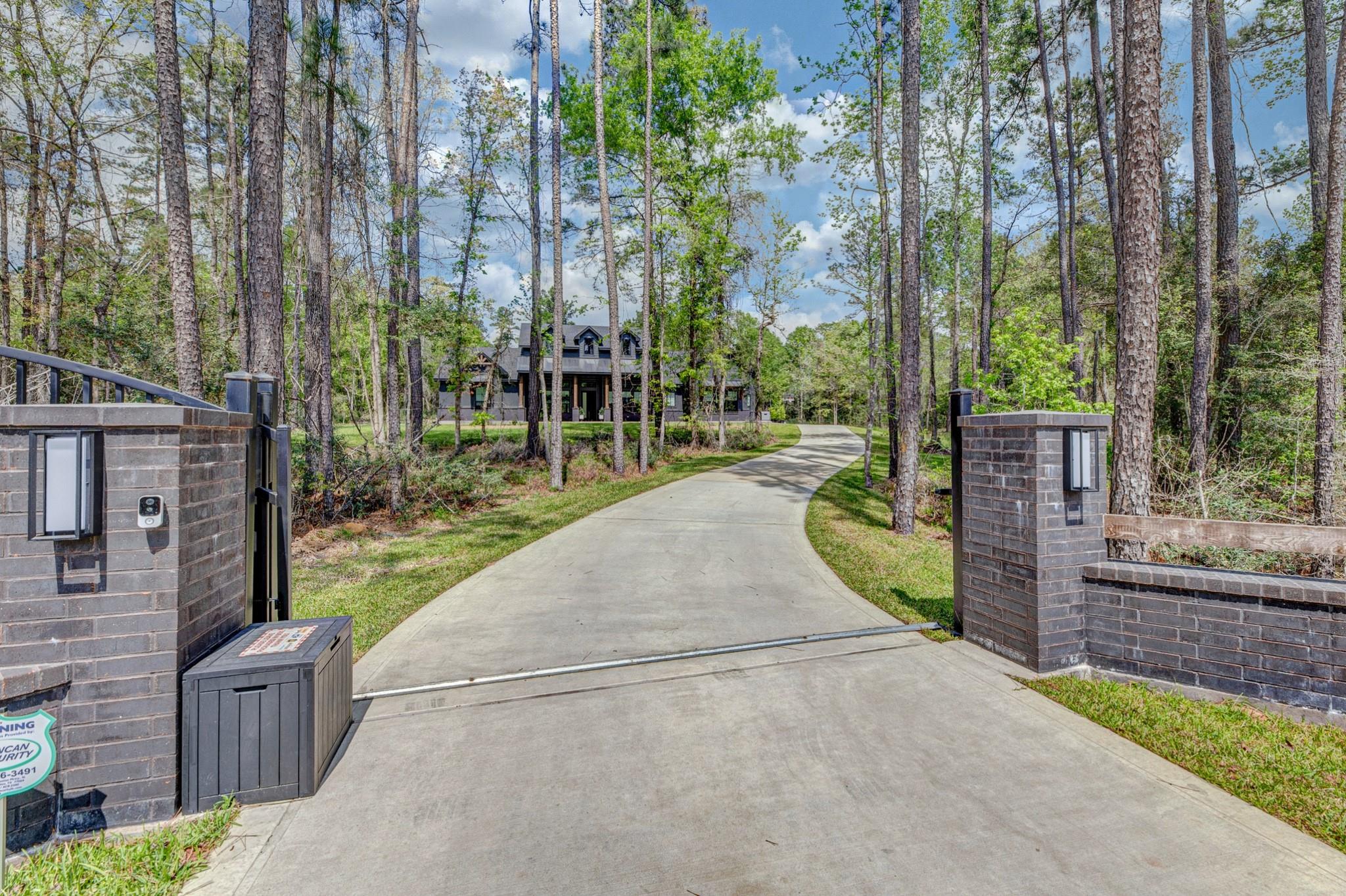 House among tall trees at the end of a curved driveway, viewed through an open brick gate.