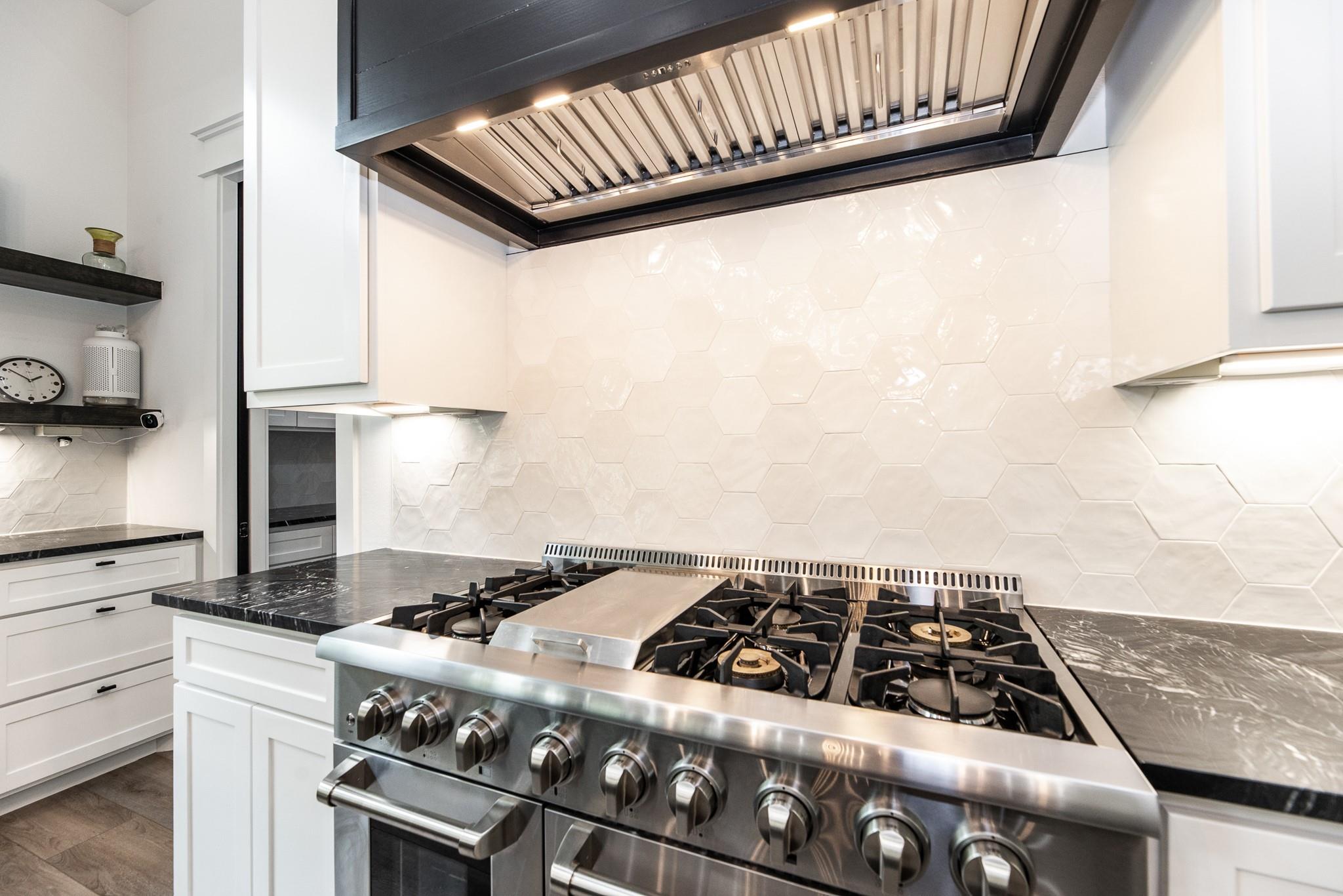 Modern kitchen with a wide stainless-steel gas range, oversized hood, and white hexagonal tile backsplash.